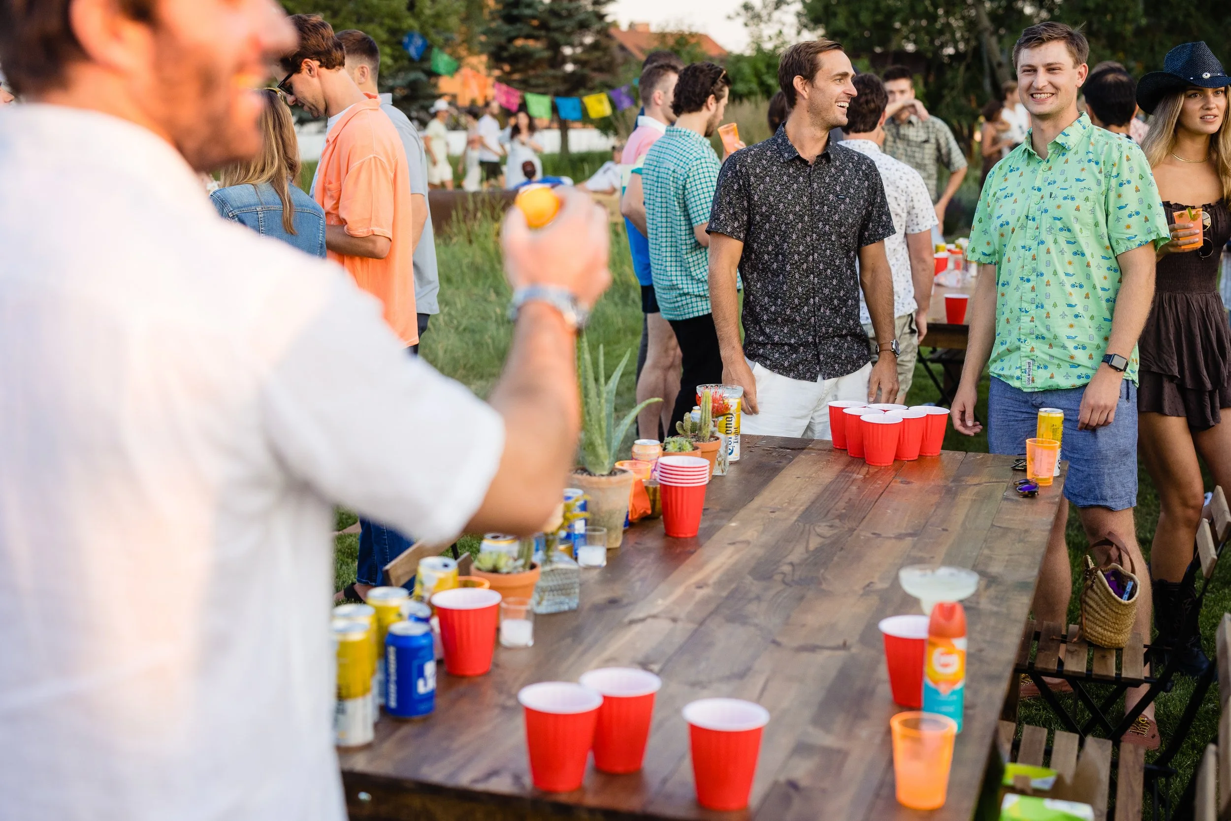 People playing beer pong outdoors during a gathering or party, with a long wooden table set up with red cups, beverage cans, and plants for decoration.