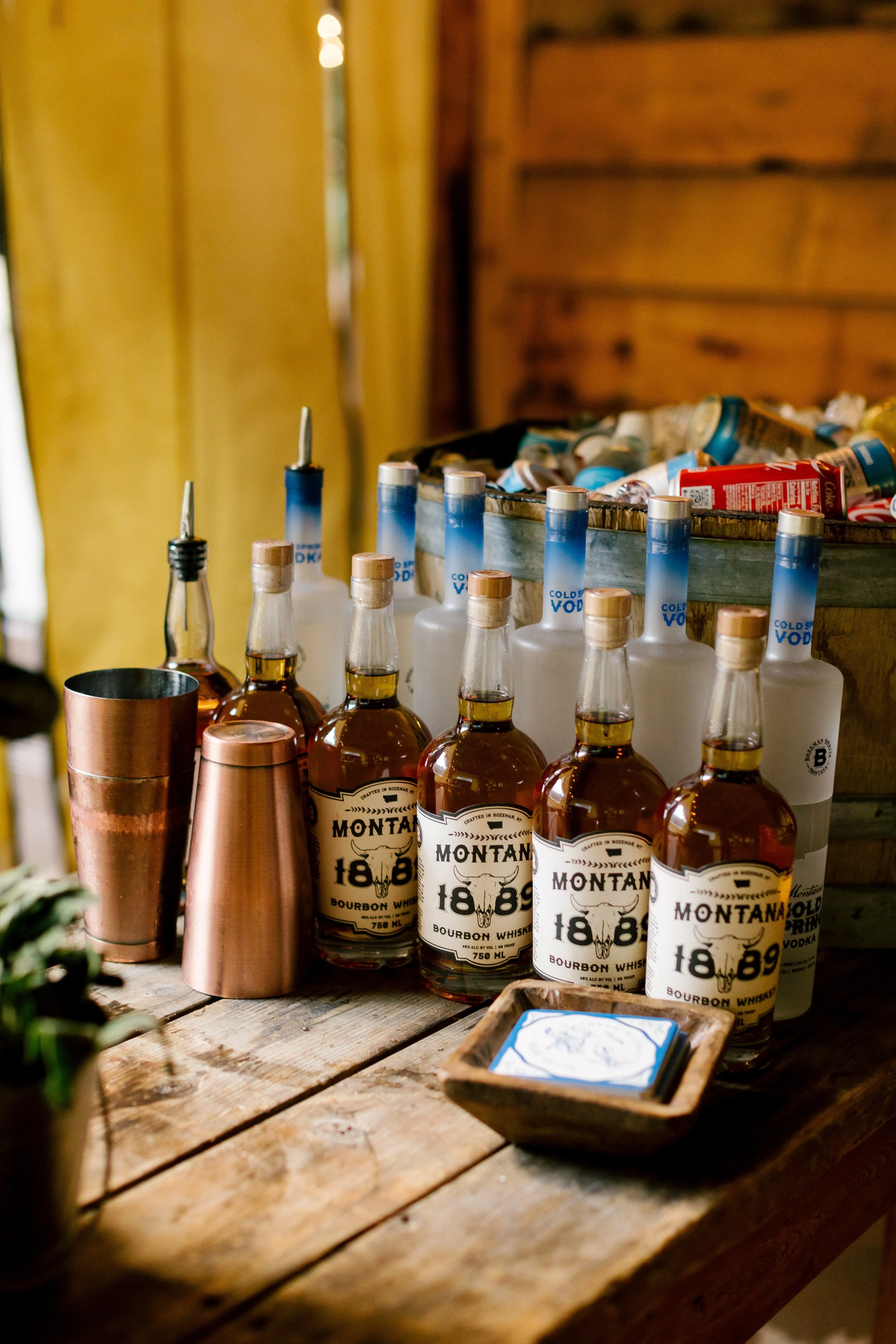 Bottles of Montana bourbon whiskey on a rustic wooden table with a copper cocktail shaker and a small wooden tray of coasters in the foreground. A wooden wall and a box filled with assorted cans and bottles are in the background.