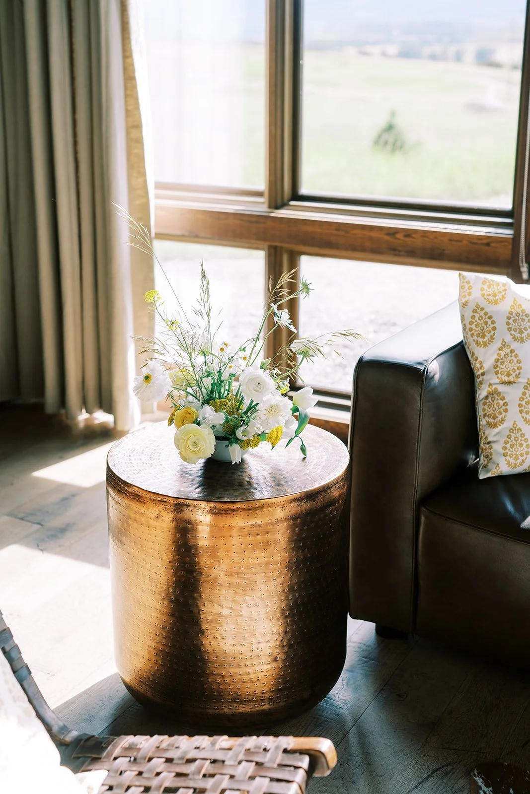 A round, textured copper side table with a white flower arrangement in a vase, next to a black leather sofa with a yellow patterned pillow, with large open windows in the background showing a grassy outdoor area.