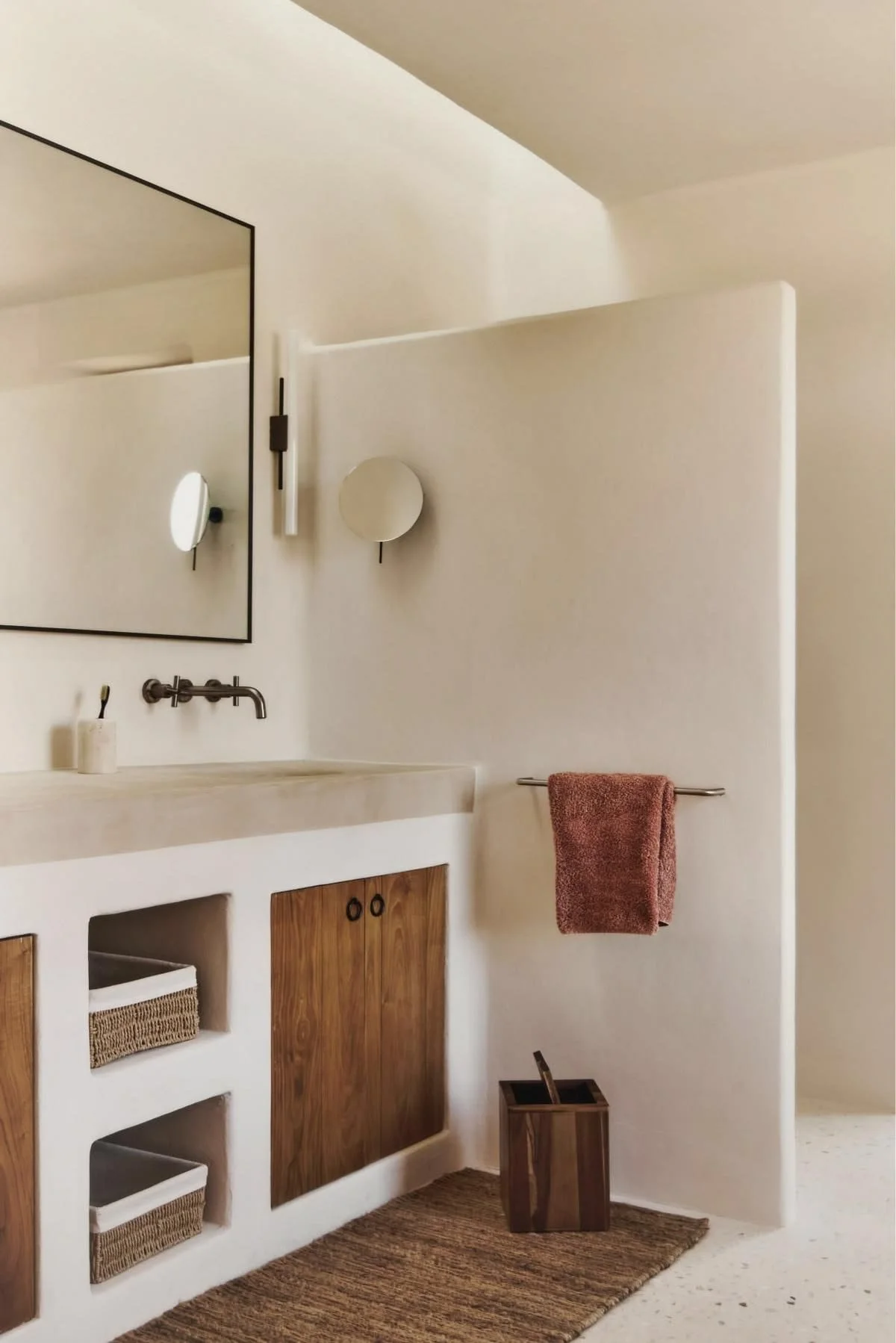 Minimalist bathroom with a mirror, a wall-mounted light fixture, two circular wall lights, a beige countertop with a built-in sink, a wooden cabinet with two doors, a red towel hanging on a towel bar, a small wooden waste bin, and a brown woven rug on the floor.