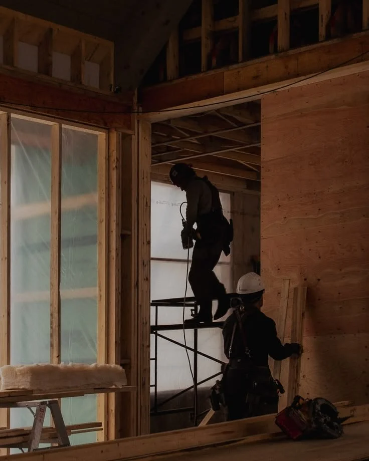 Two construction workers in safety gear working inside a building under construction, with exposed wooden framing and scaffolding.