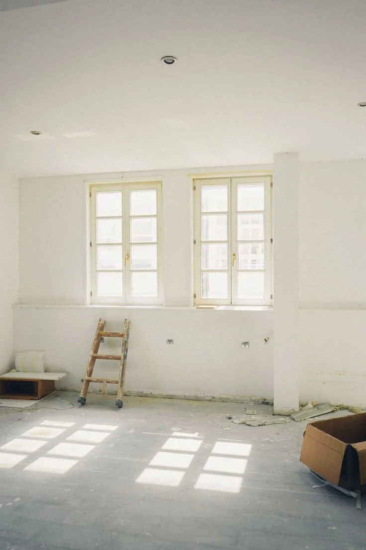 Empty room with large windows, construction tools, and debris on the floor, indicating ongoing renovation.