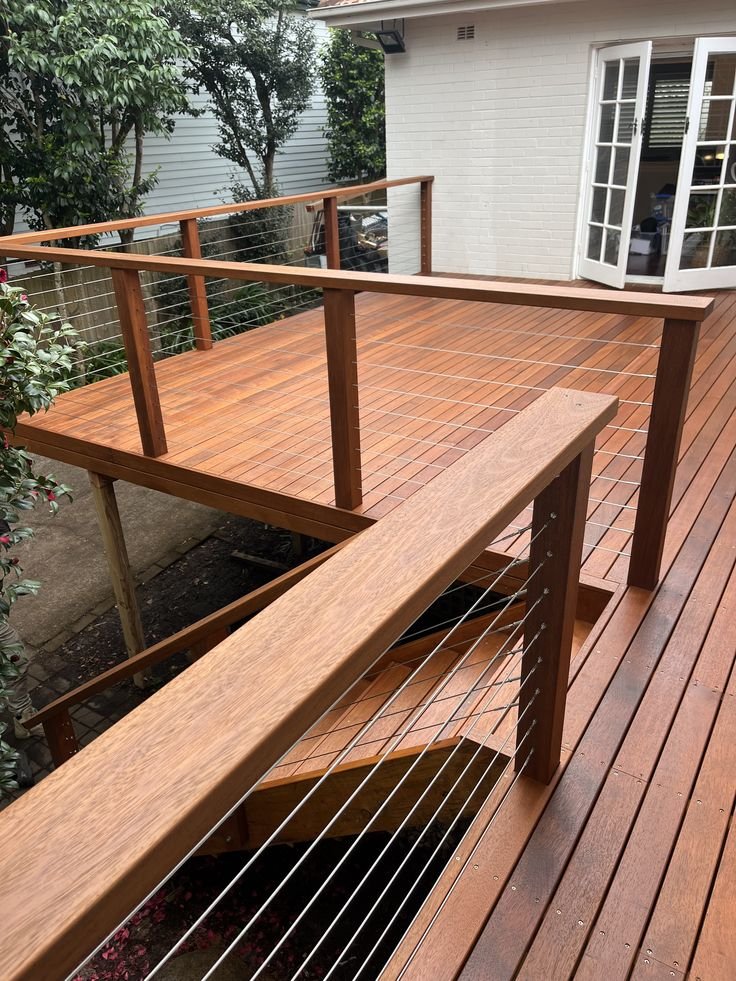 Wooden outdoor deck with staircase and metal wire railings, adjacent to a white house with glass-paneled doors.