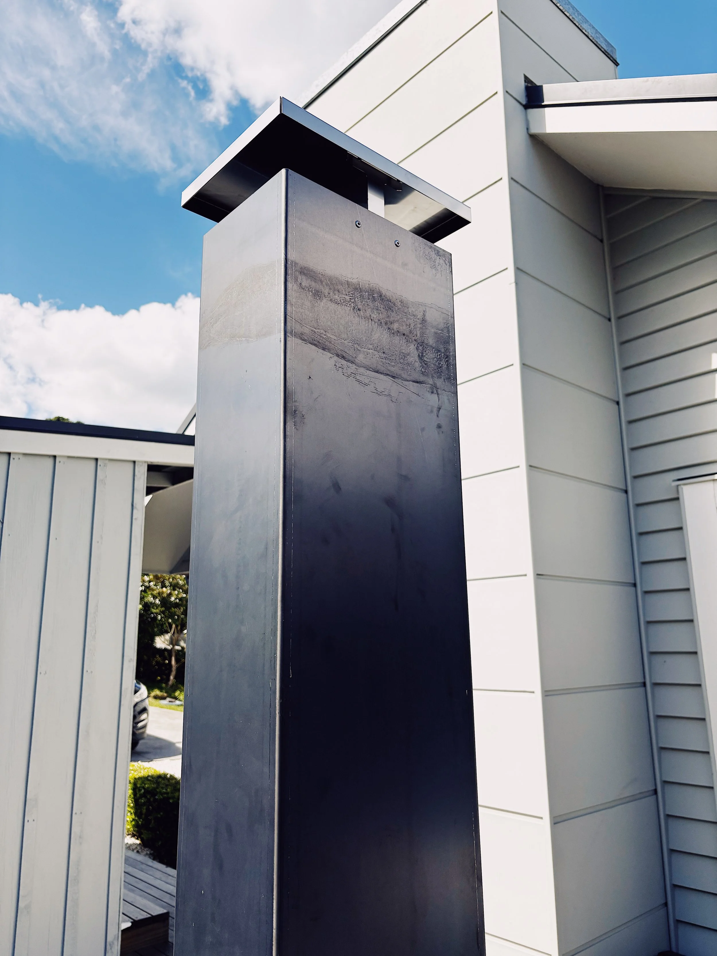 Modern black outdoor mailbox on a metal post outside a house under a blue sky with some clouds.