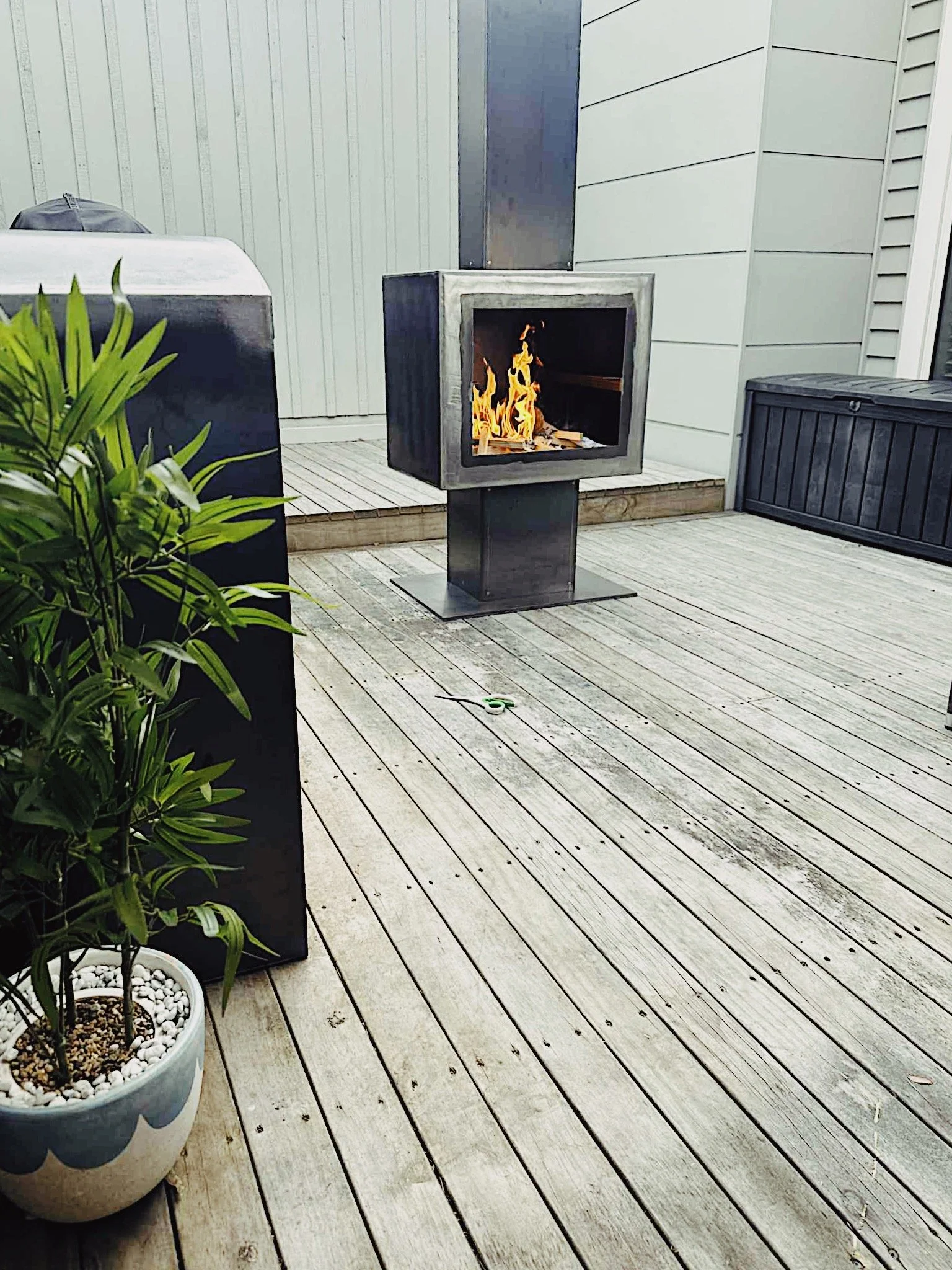 A modern outdoor patio with a metal fire pit, potted plant, and storage boxes.
