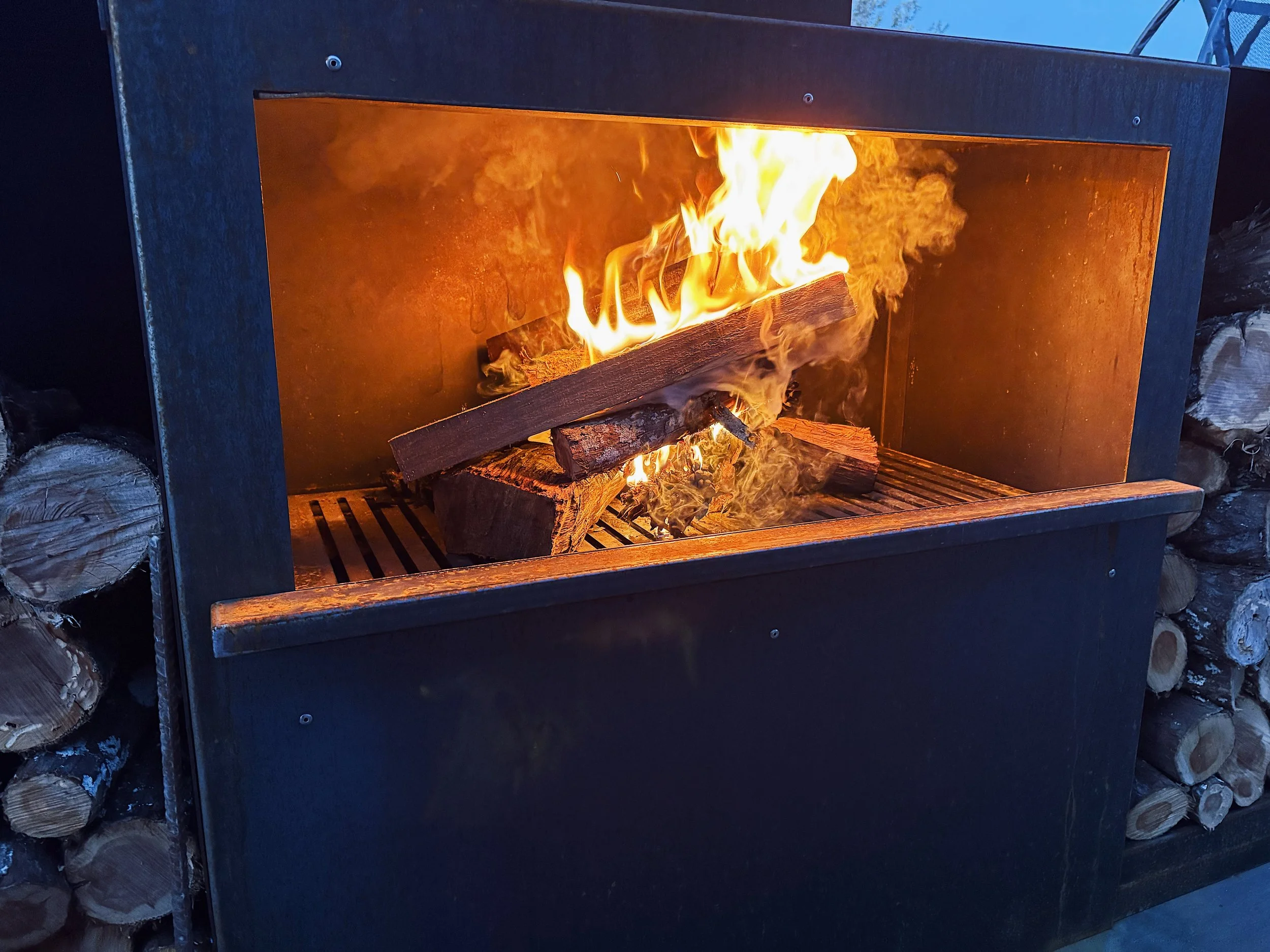 A modern outdoor fireplace with flames burning and logs inside, surrounding a stack of firewood.