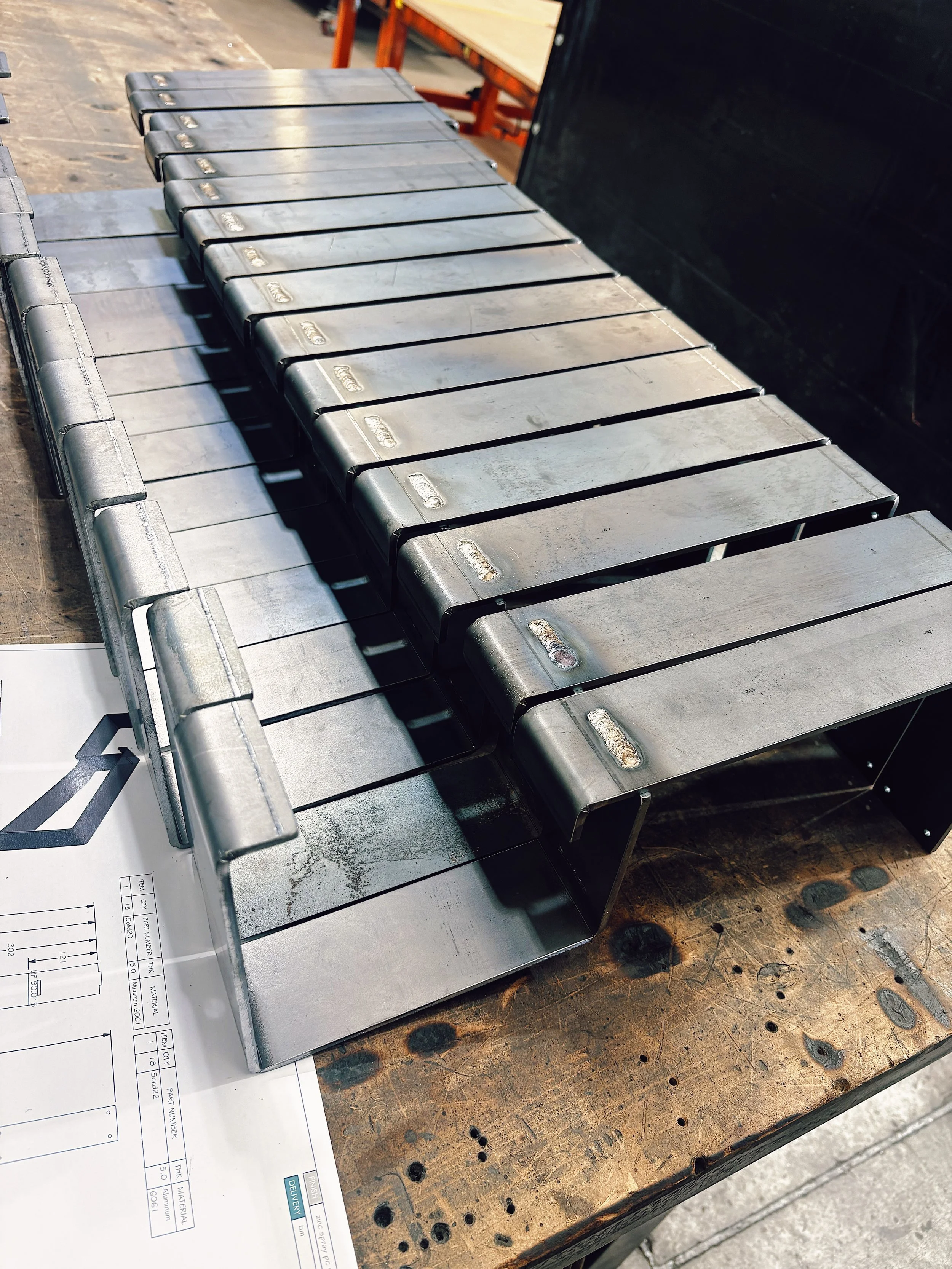 Metal working project with bent metal pieces on a workbench, blueprints in the corner.