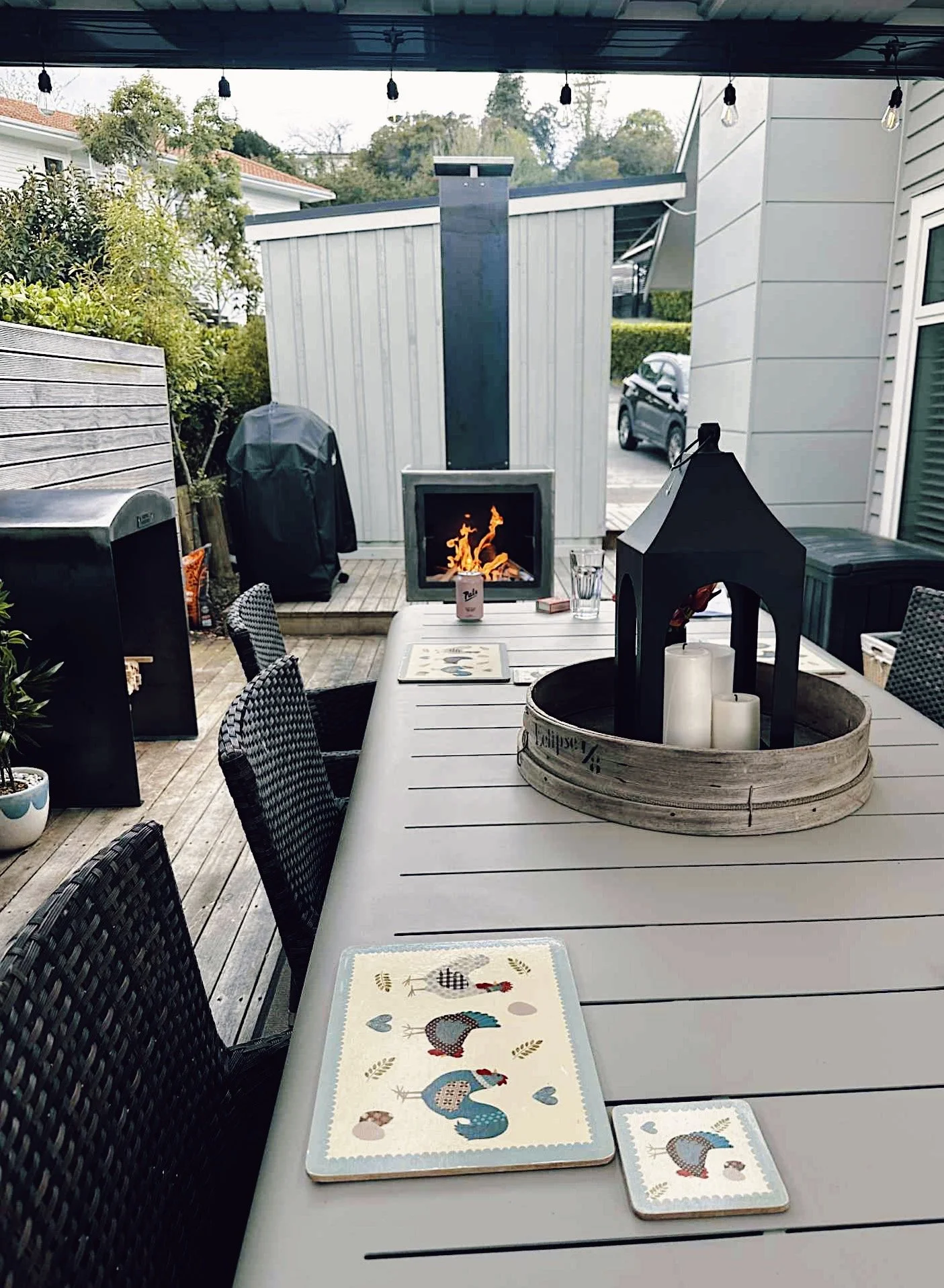 Outdoor patio with a white table, chairs, a fireplace with a visible flame, and decorative candles in a black lantern on a tray. Boards and outdoor storage units, a barbecue covered with a black cover, potted plants, and string lights overhead.