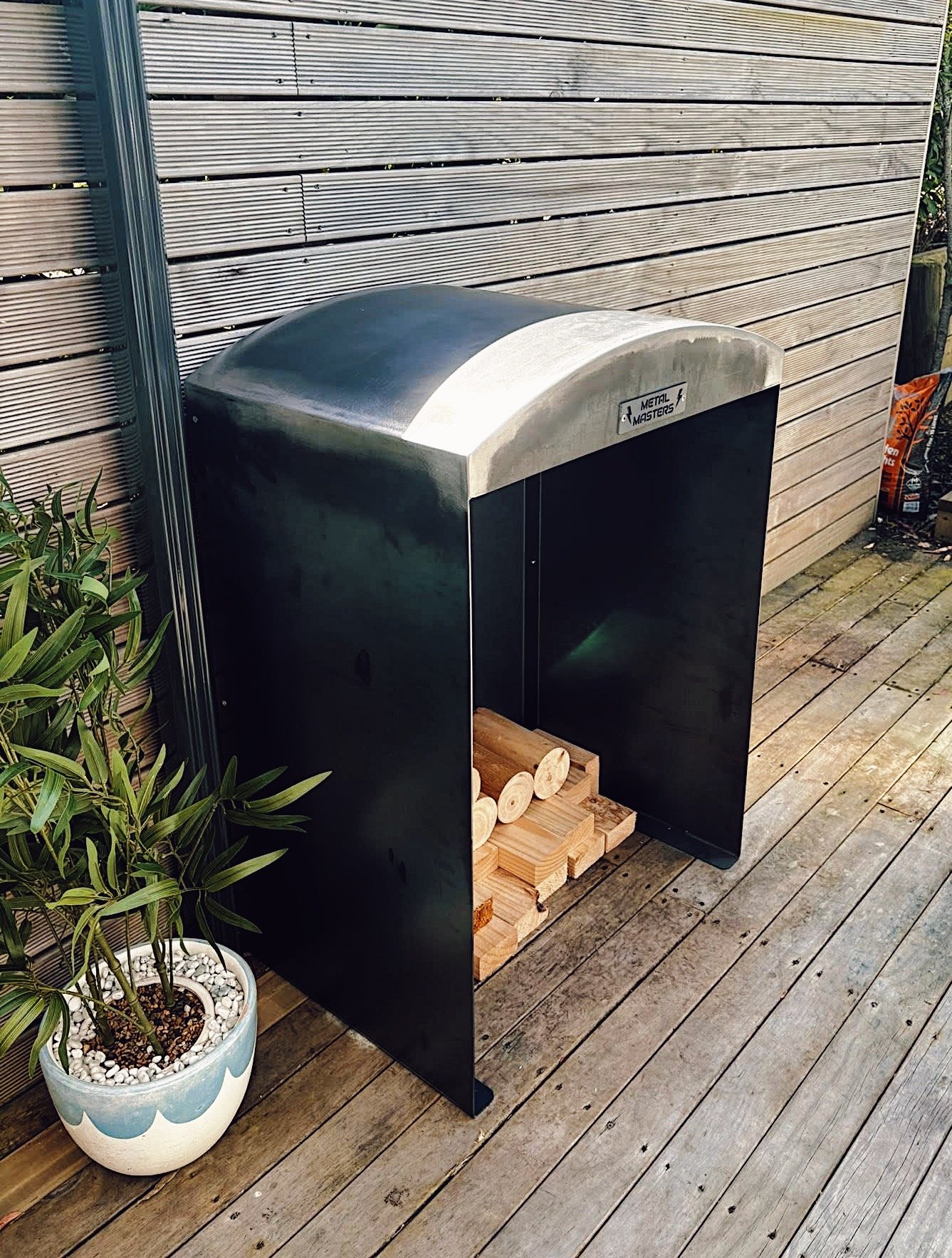 A metal firewood storage box with a curved top, filled with cut firewood, located on a wooden deck next to a potted plant with green leaves.