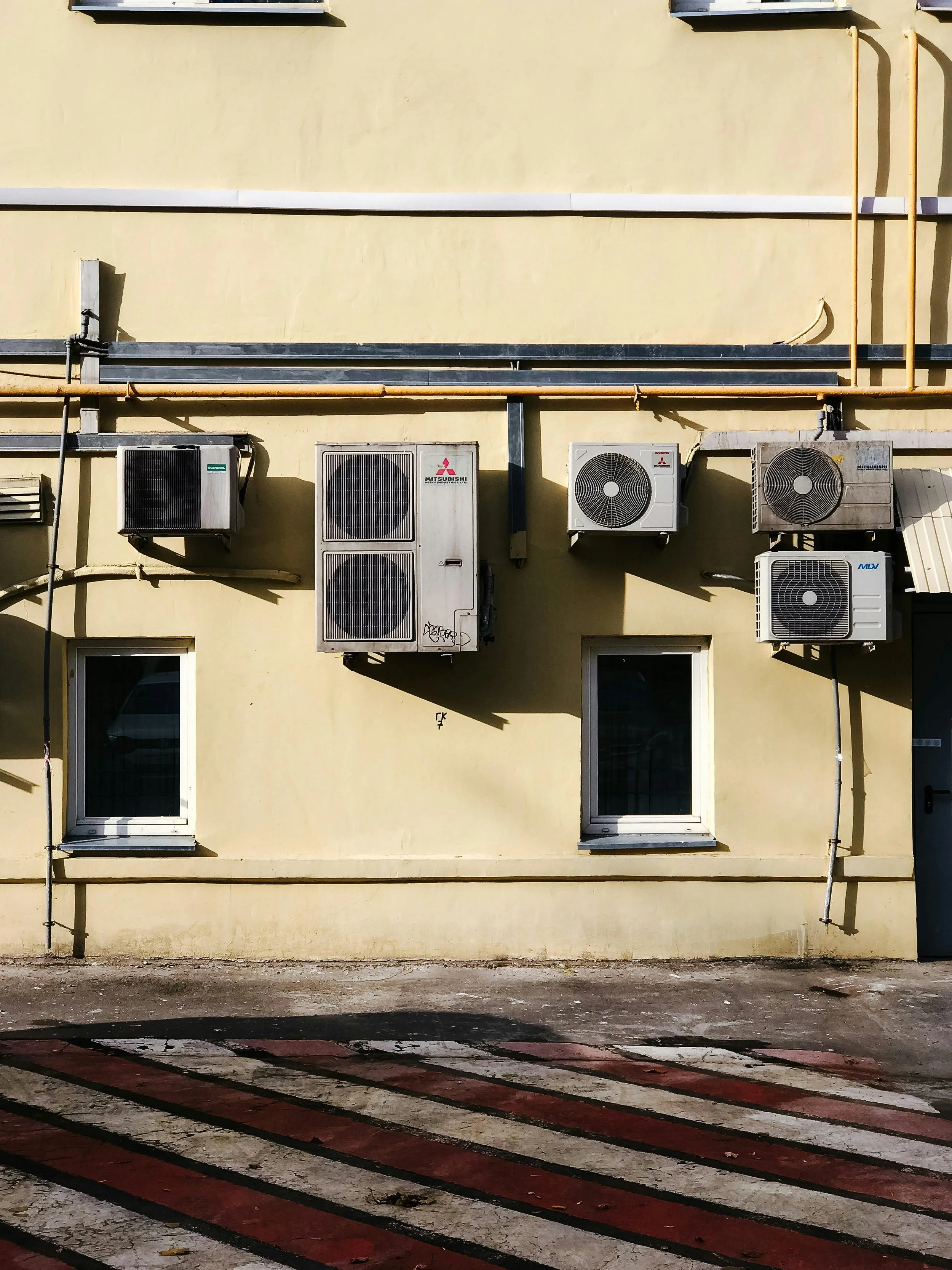 Façade d'un bâtiment jaune avec deux fenêtres et plusieurs unités de climatisation sur le mur, avec un passage piéton en premier plan.