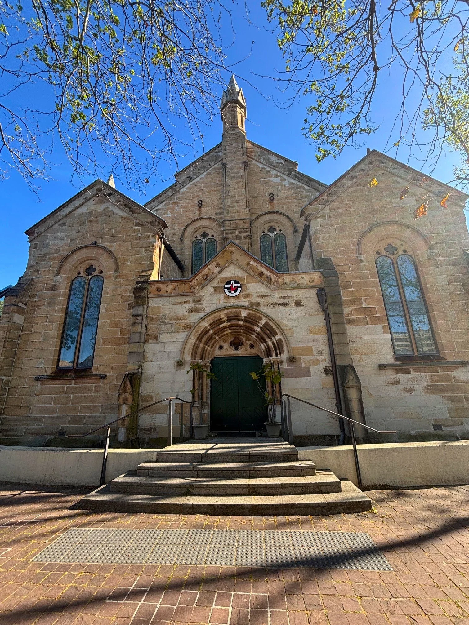 Front view of a stone church with steps leading up to the entrance, surrounded by trees with some leaves fallen, clear blue sky above.