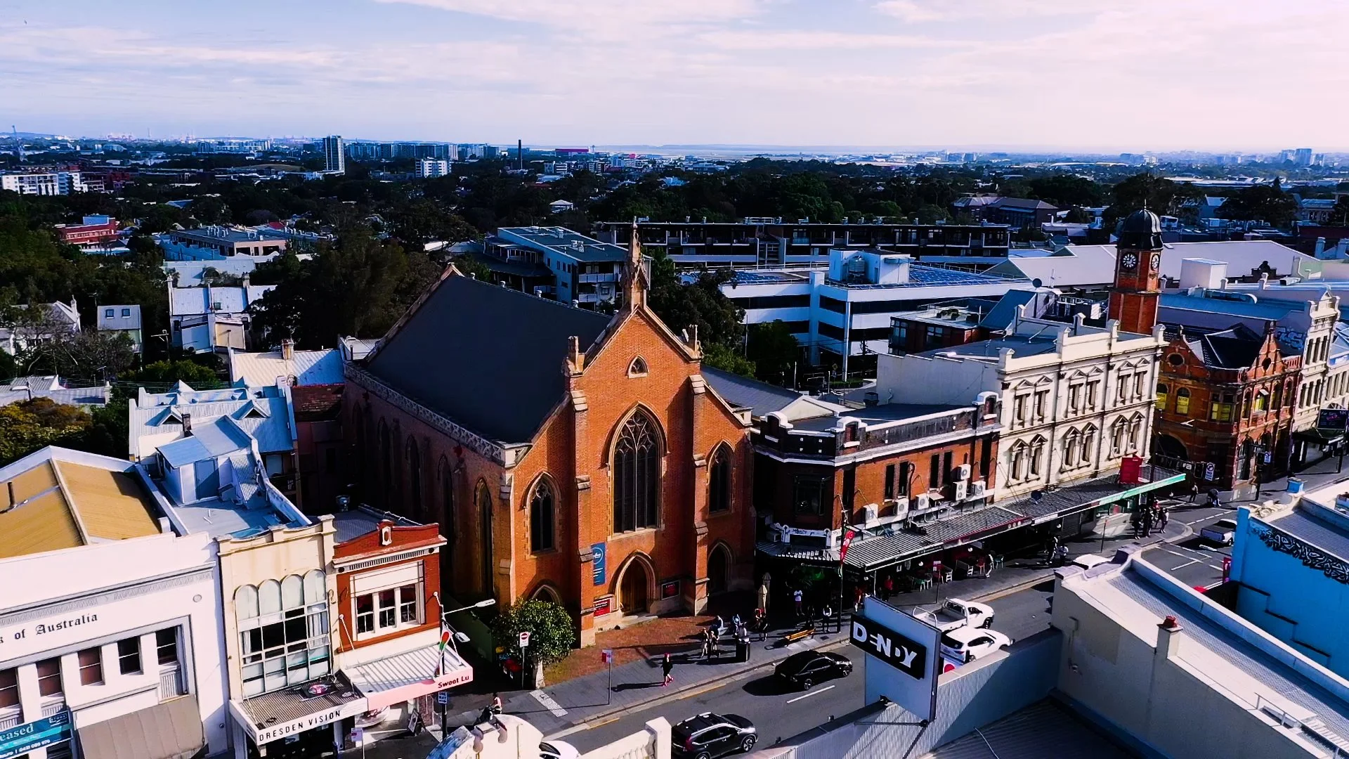 A cityscape featuring a historic red brick church with gothic windows, modern buildings, and trees under a partly cloudy sky, with cars and pedestrians on the street below.
