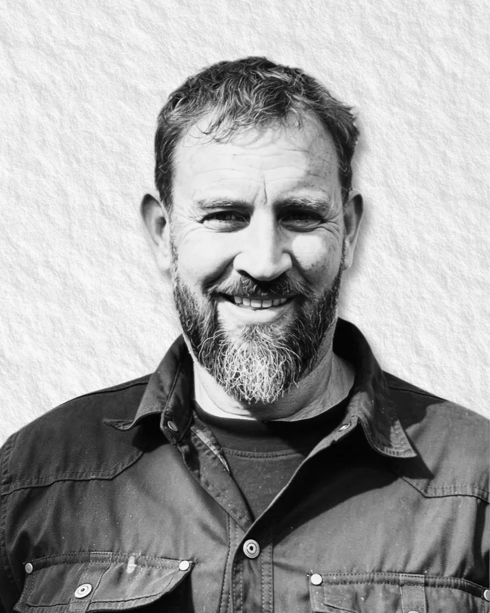 Black and white portrait of a man with a beard, smiling, wearing a button-up shirt over a t-shirt, standing against a light-colored textured background.