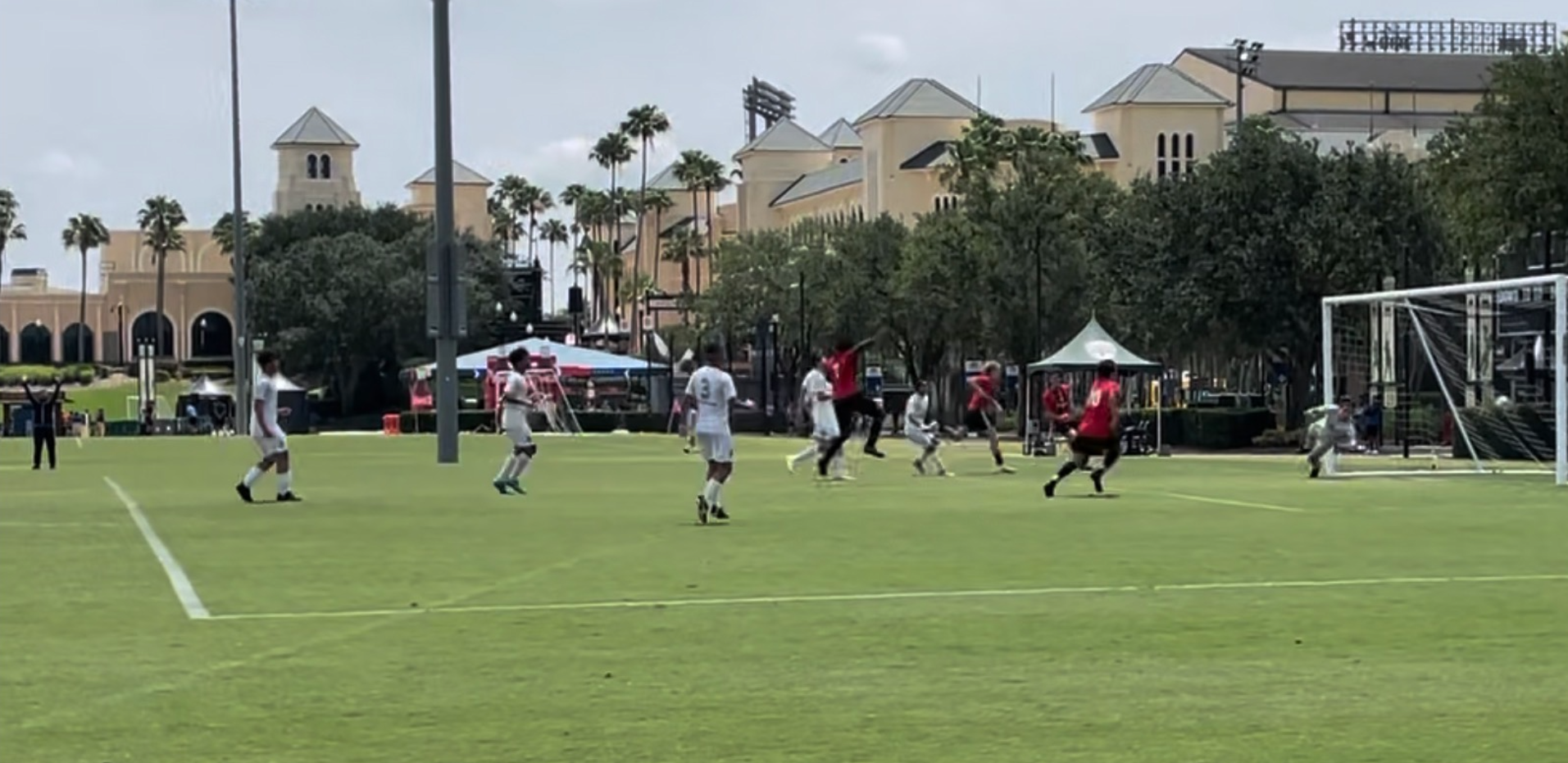 Youth soccer game on a green field with players in white and red uniforms, a goalkeeper in gray, and a large stadium in the background.