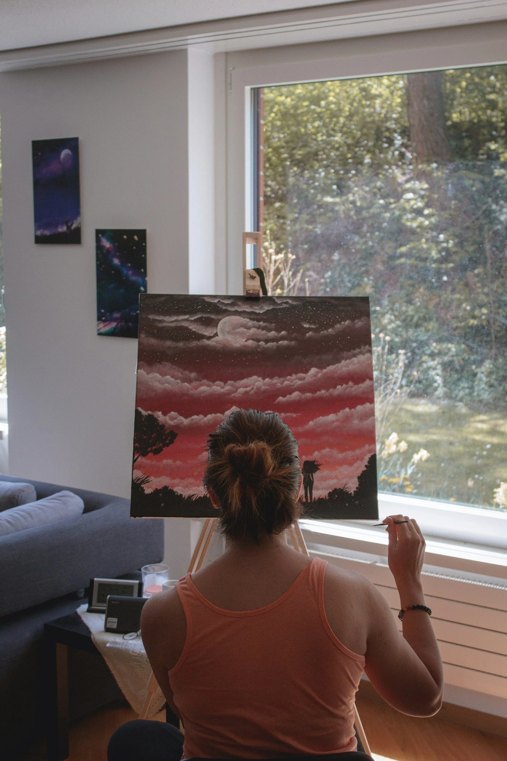 Woman painting a landscape scene featuring a moon, clouds, and a person with a dog in a room with large window, space-themed artwork on walls, and a table with picture frames.
