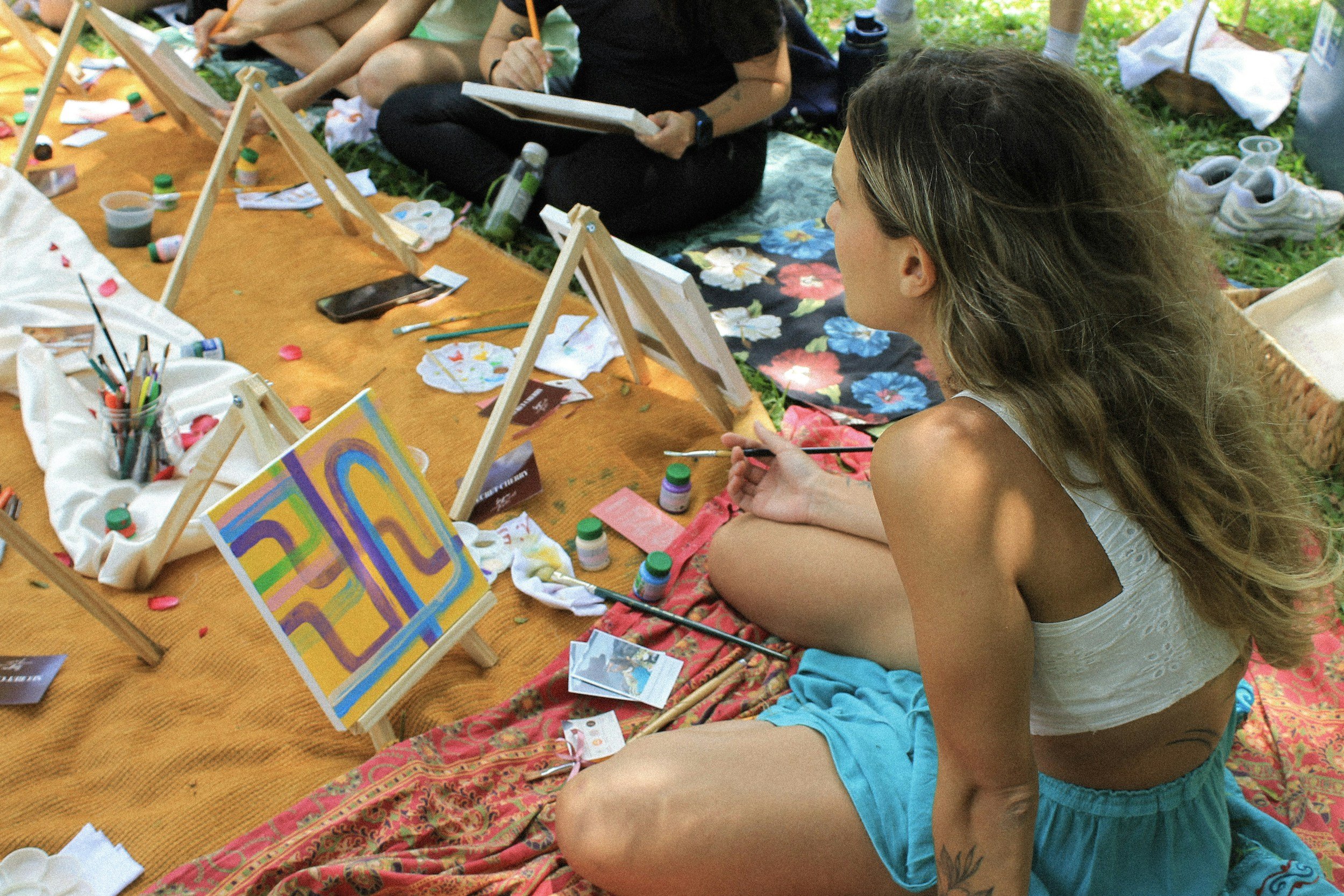 Woman with long wavy hair painting on a canvas at an outdoor art event. She is seated on a floral blanket surrounded by art supplies, including paints, brushes, and small canvases. Other people can be seen in the background also engaging in painting activities.