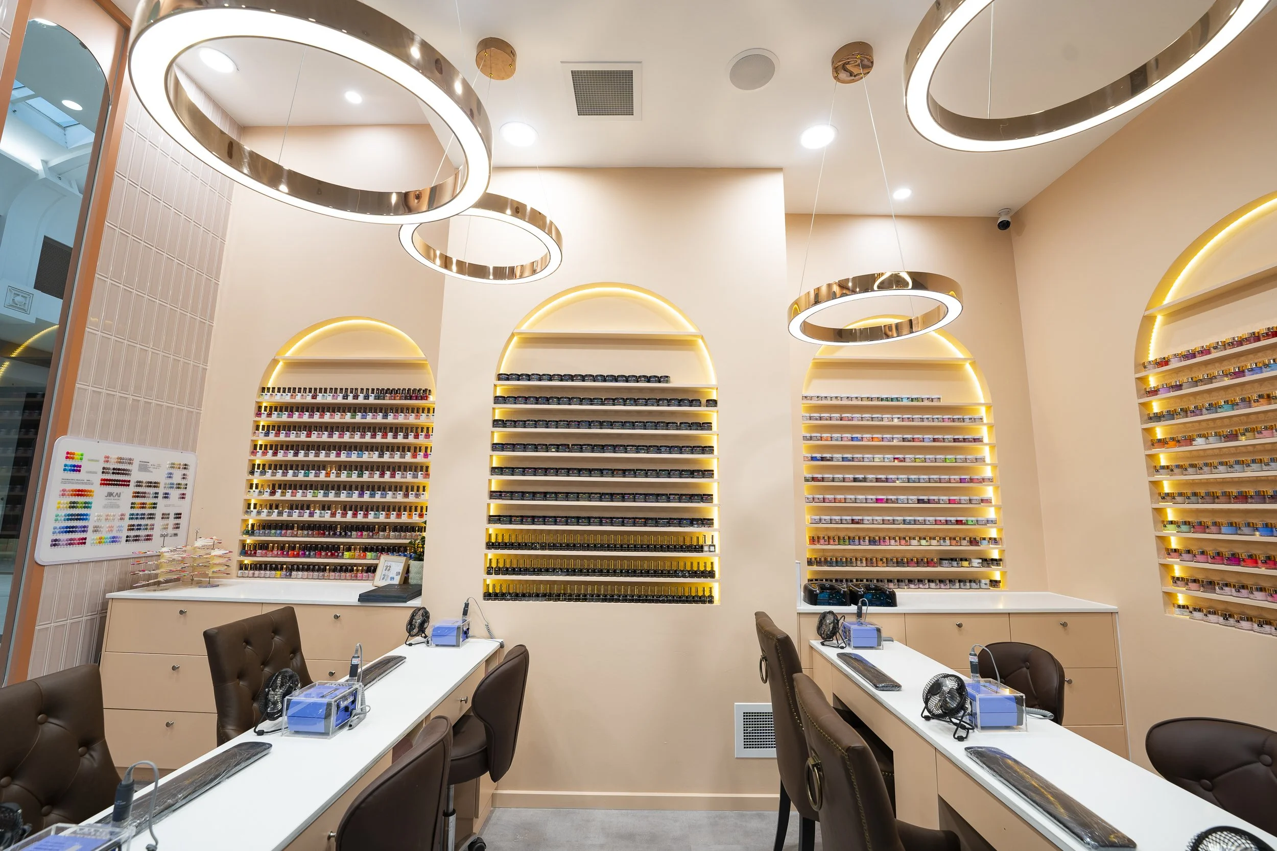 Interior of a nail salon with nail polish shelves on the walls, tables with chairs, manicure tools, and modern lighting fixtures.