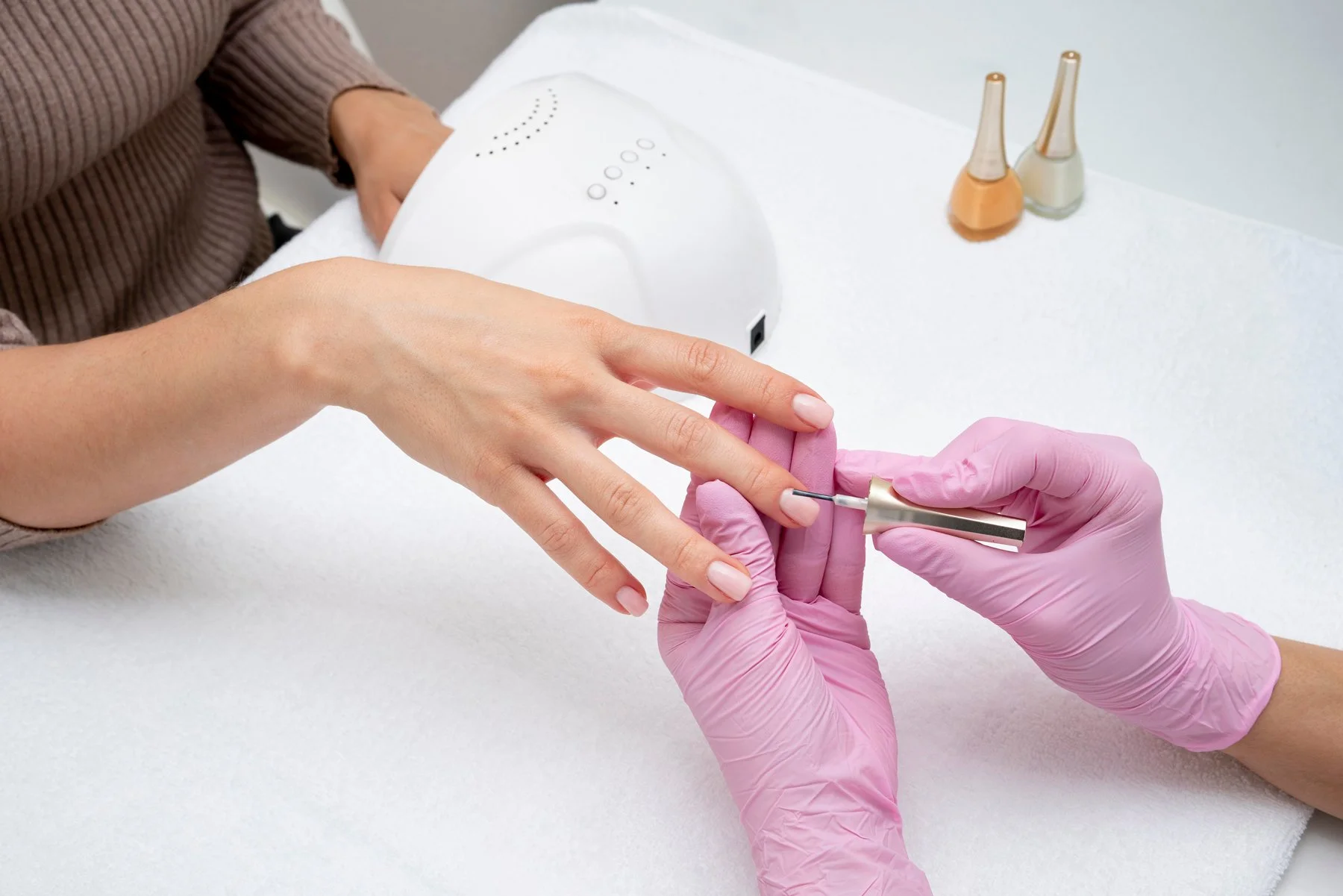 Close-up of a woman getting a manicure with a pink-gloved technician filing her nails at a salon, with manicure tools and nail polish bottles on the table in the background.
