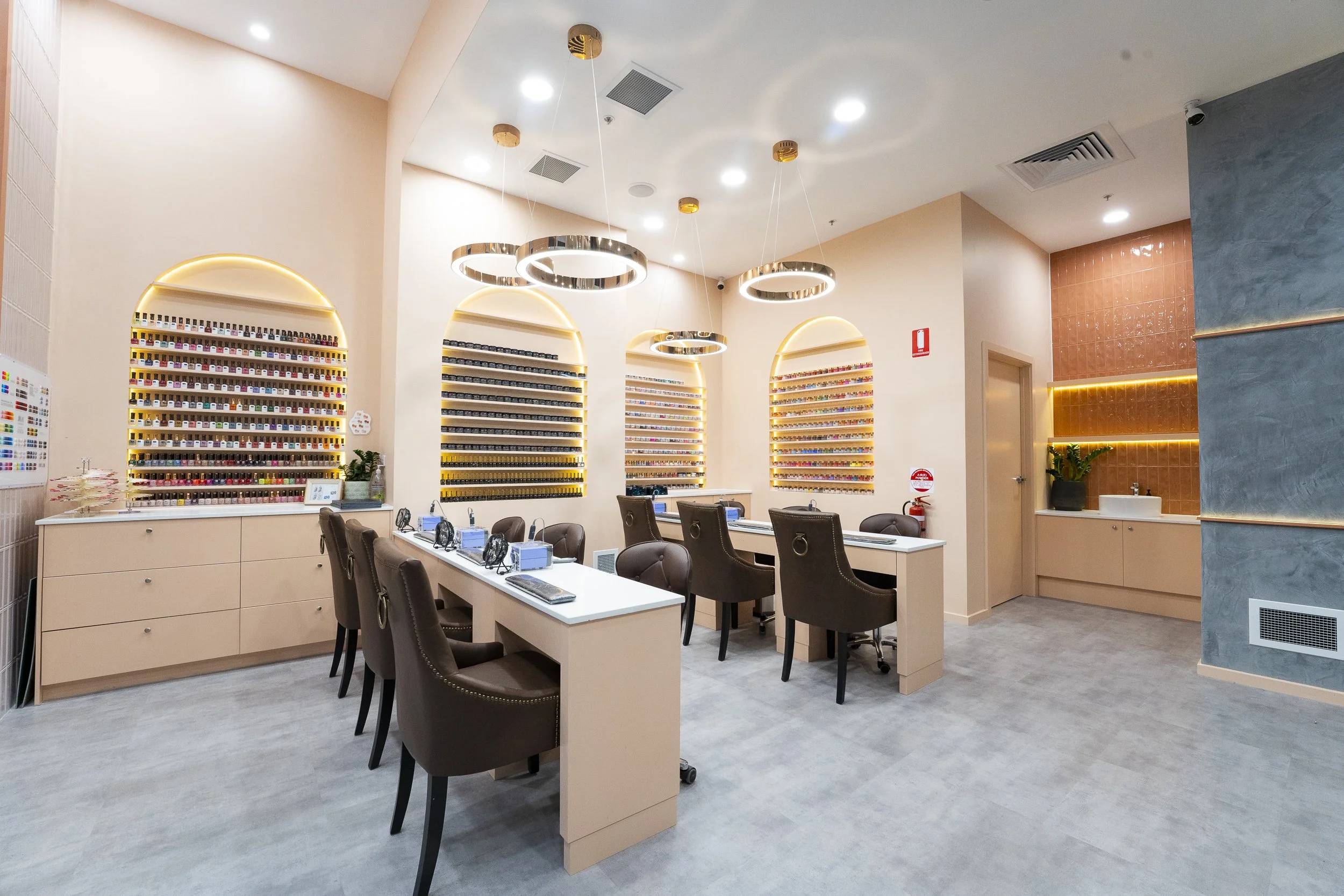 Nail salon interior with four workstations, each equipped with a chair, a lamp, and nail polish display shelves on the wall. There are additional shelves with lights, a small sink area, and various nail polish bottles on display. The atmosphere is modern and well-lit.