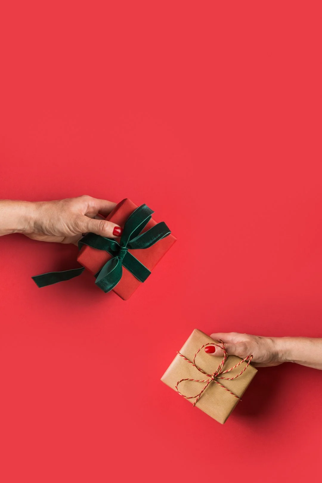 Two hands holding wrapped Christmas gifts against a red background, one with a green ribbon and the other with red twine.