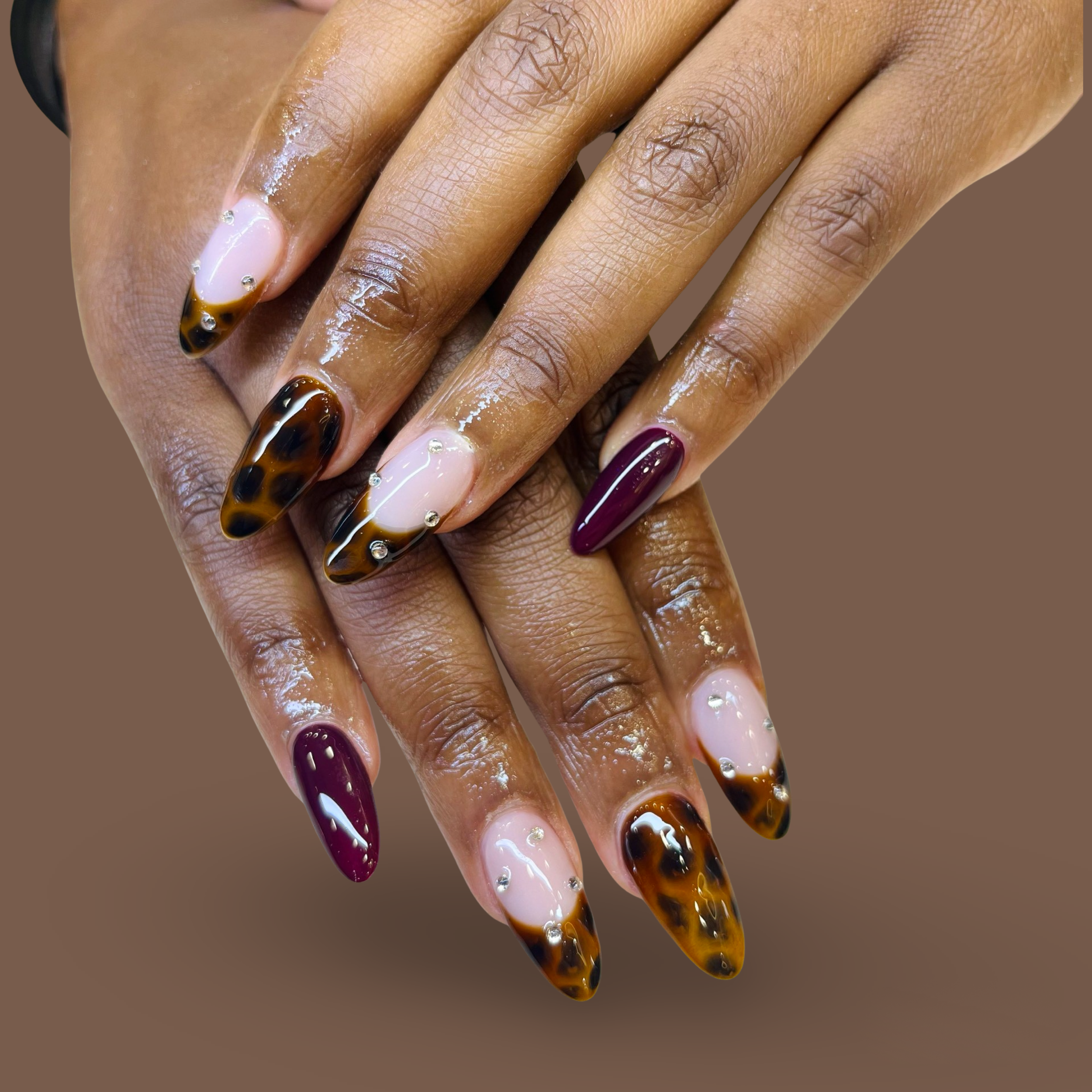 Close-up of hands with decorated acrylic nails, featuring a tortoise shell and maroon color scheme, with some nail tips adorned with small rhinestones.
