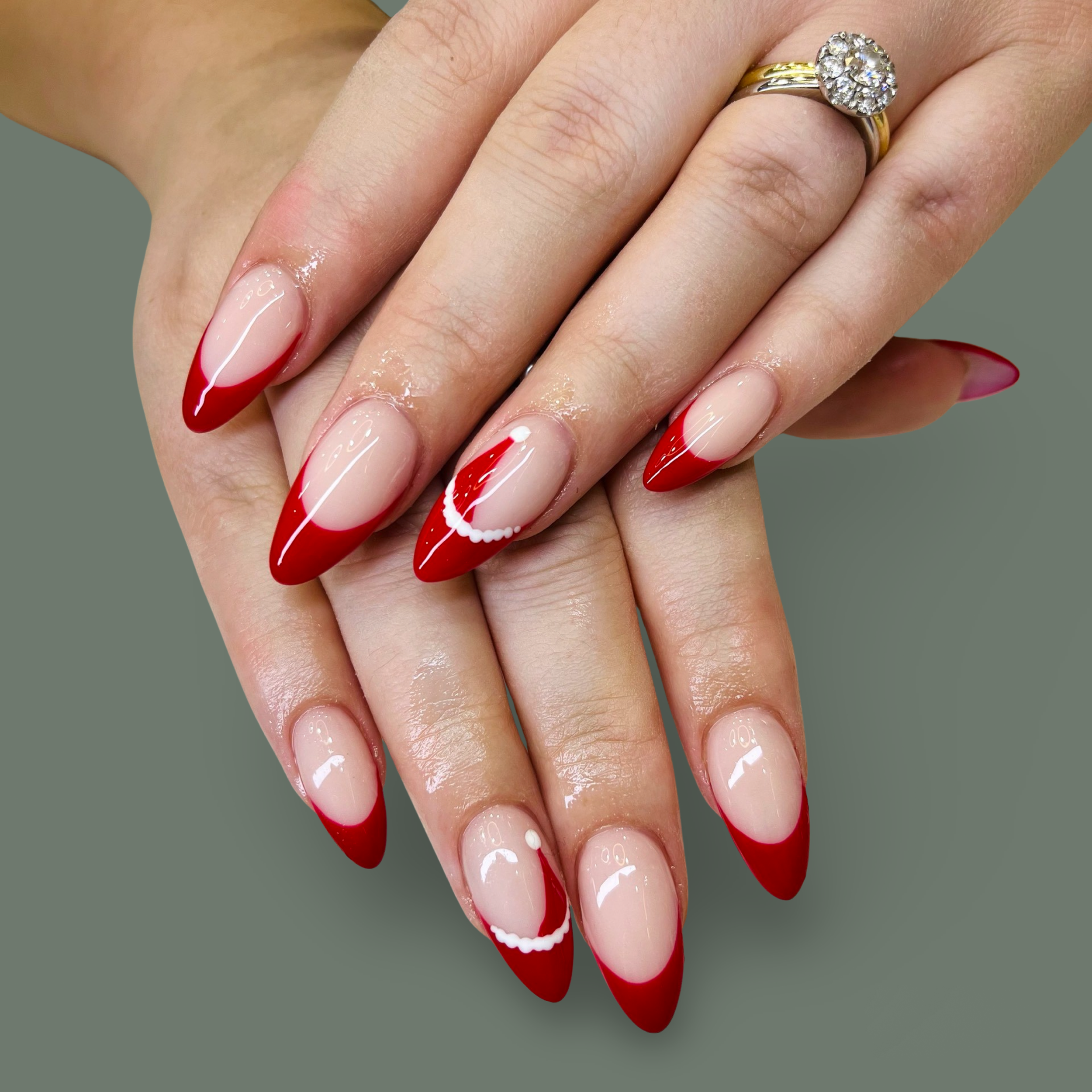 Manicured nails with red French tips and white accents on a woman's hands, with a gold ring featuring a cluster of diamonds.