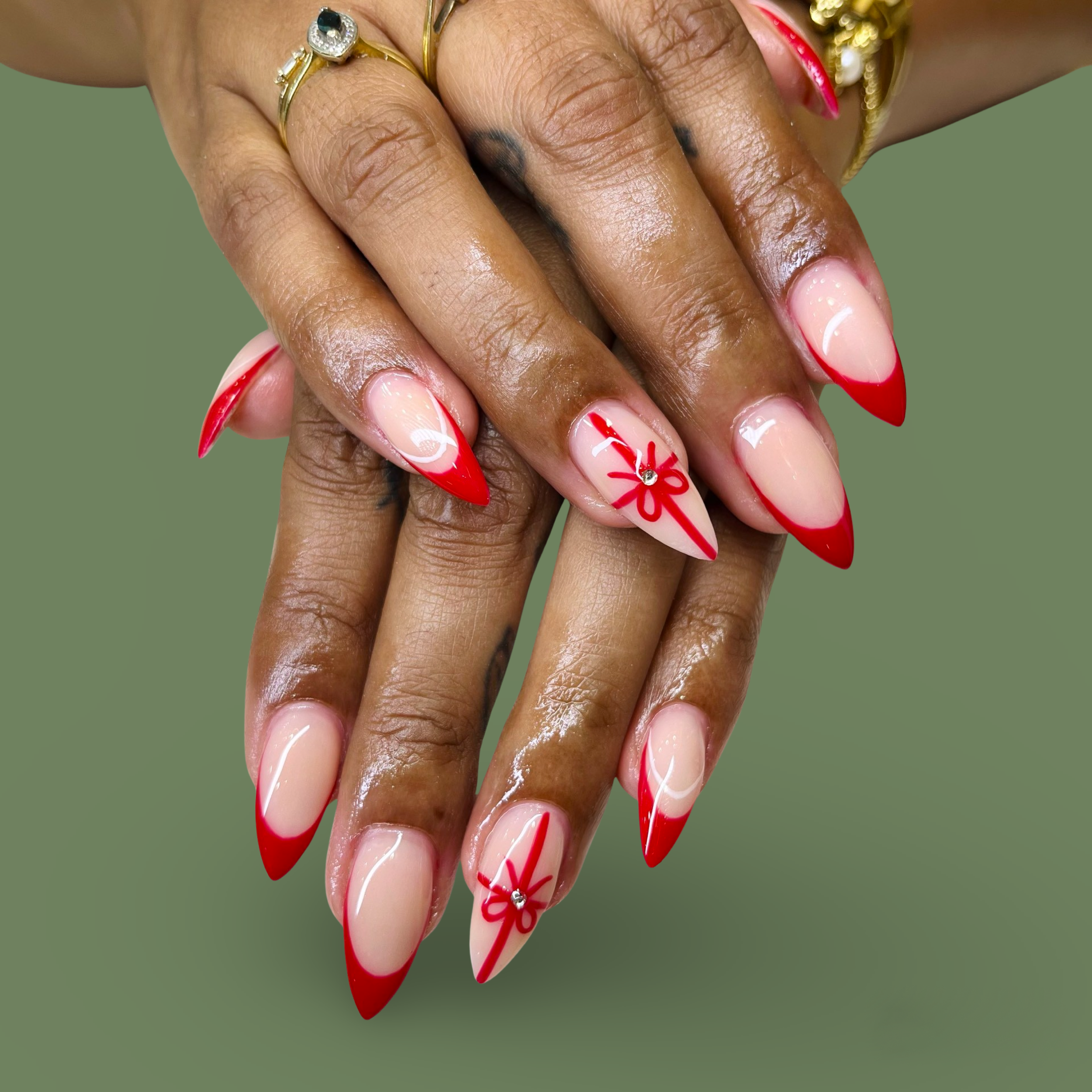 Close-up of hands with manicured nails featuring red tips and a festive bow design on two accent nails, with gold rings and jewelry.