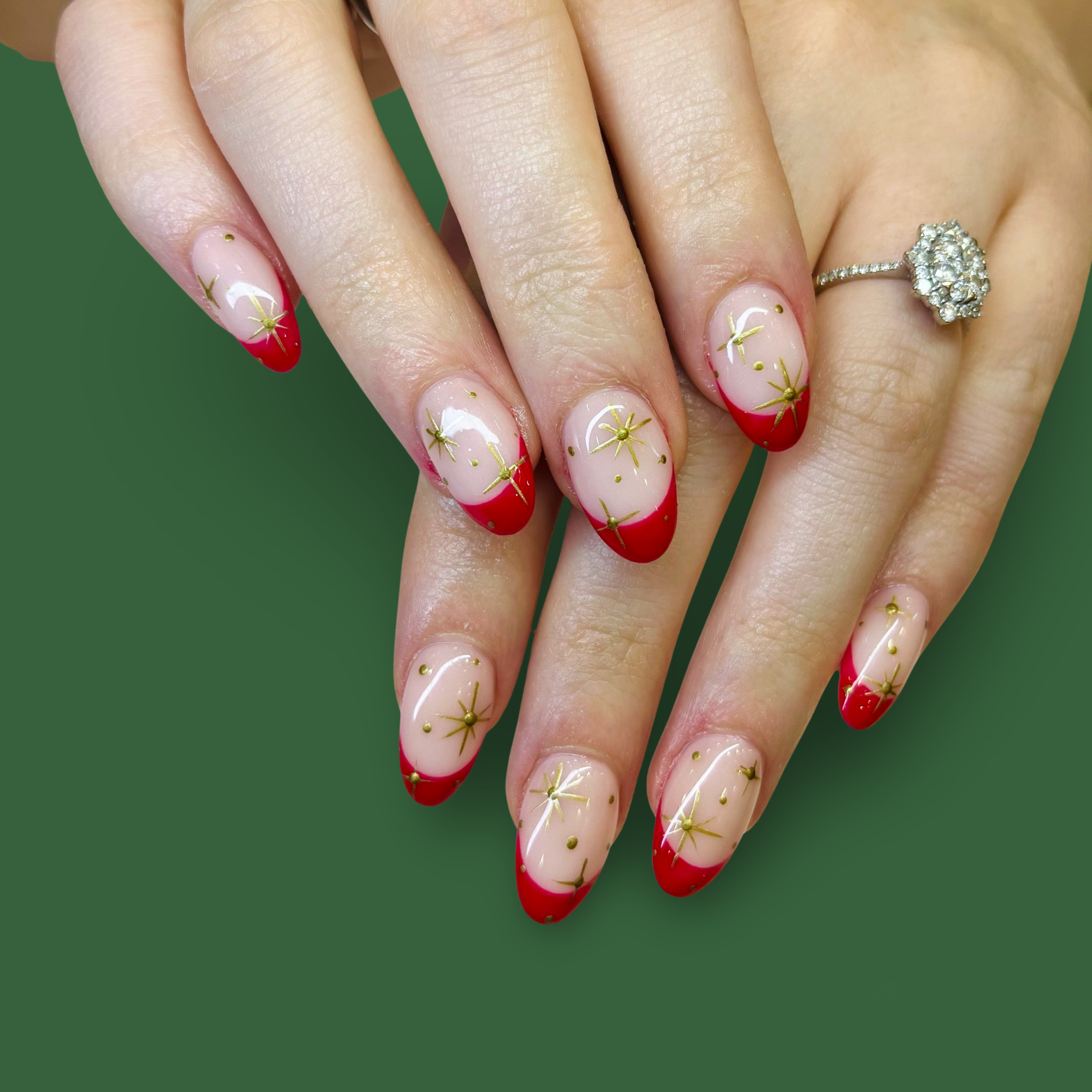 Hand with decorated nails featuring a red and gold starburst design, a diamond ring on the ring finger, and a green background.