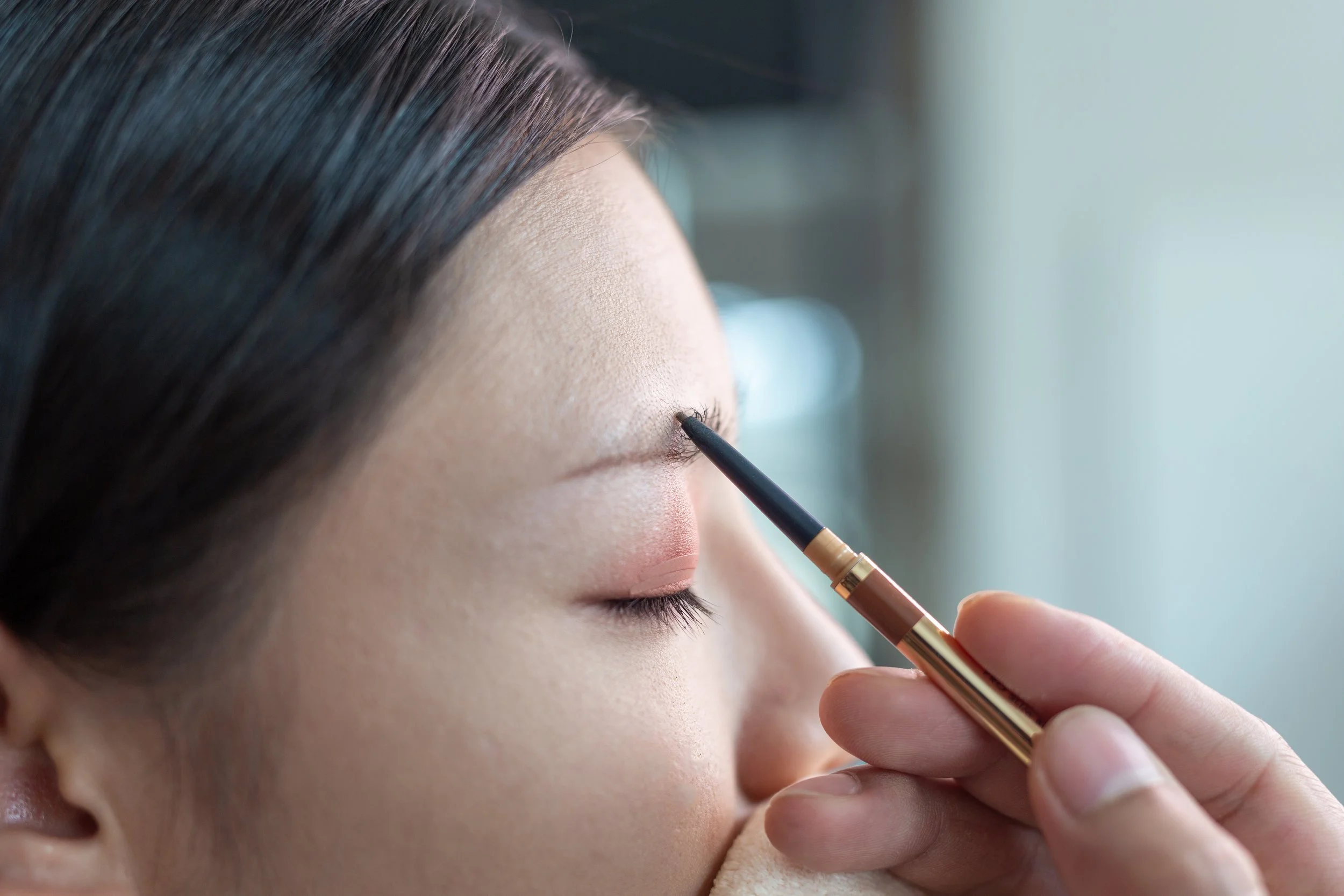 Close-up of a woman with her eyes closed as a makeup artist applies eyebrow makeup with a thin brush.