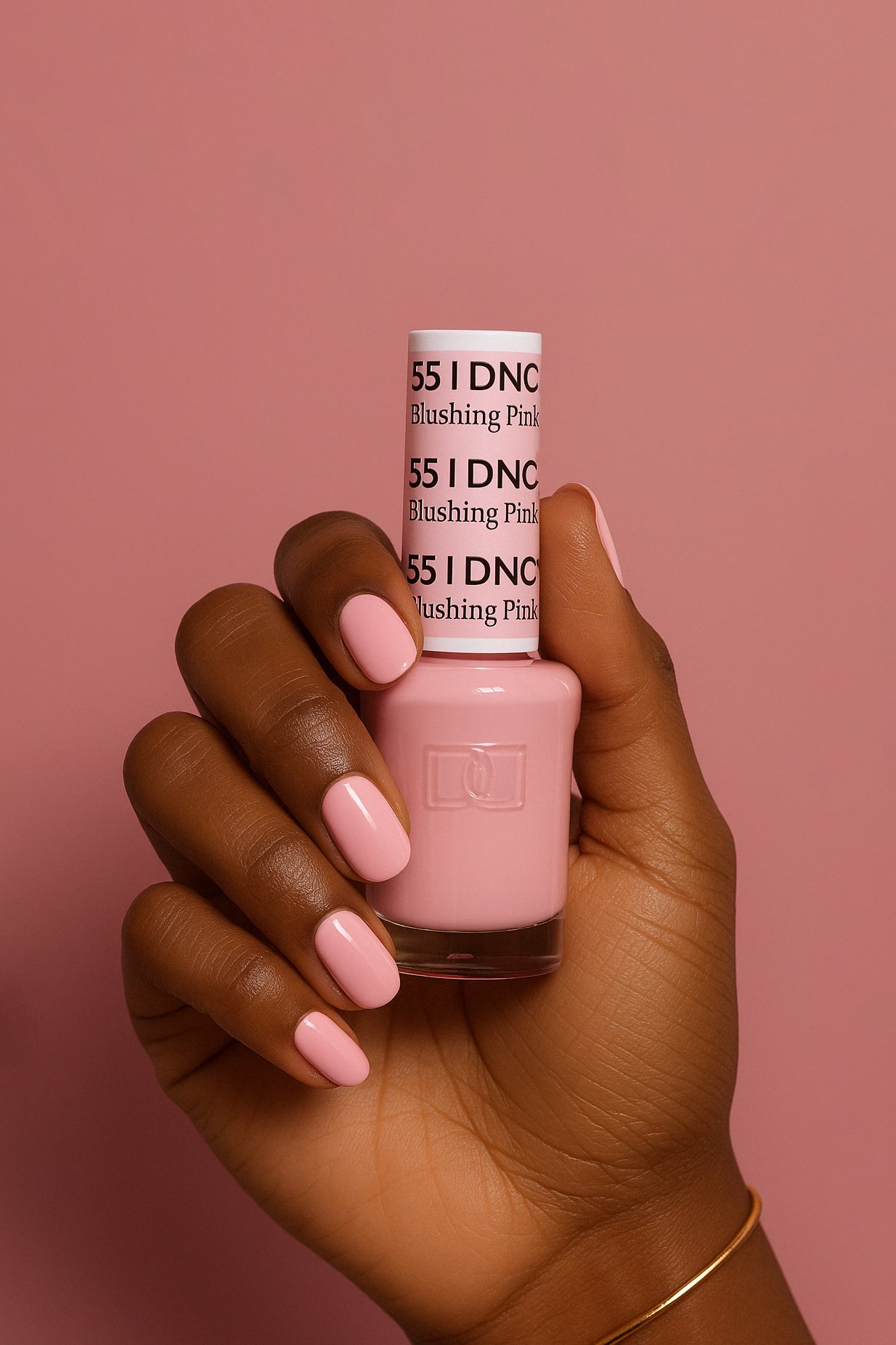 Hand with painted pink nails holding a pink nail polish bottle labeled "55 I DNC Blushing Pink" against a pink background.