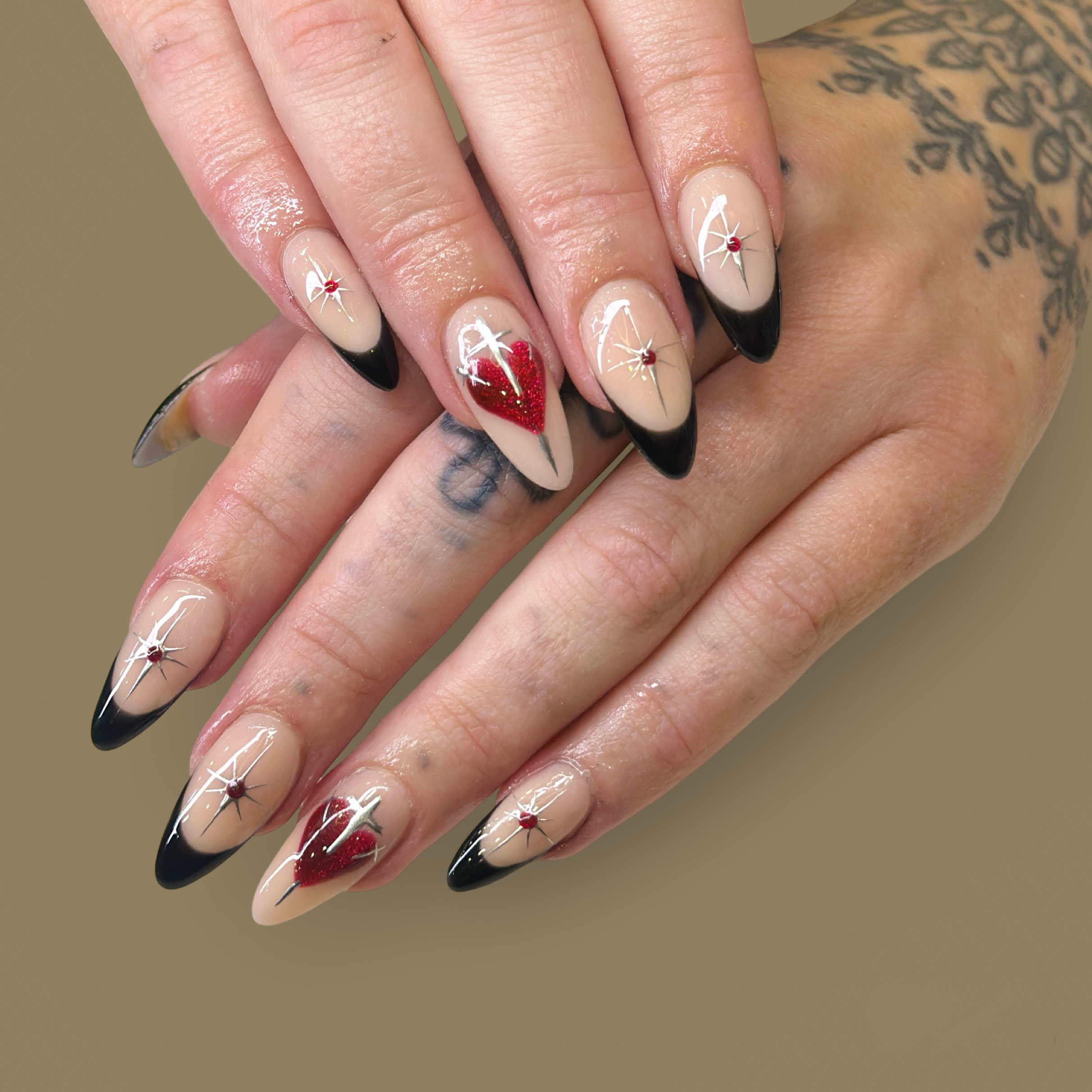 Close-up of hands with long, almond-shaped nails featuring beige base color, black tips, red heart, and starburst designs with small red gems.