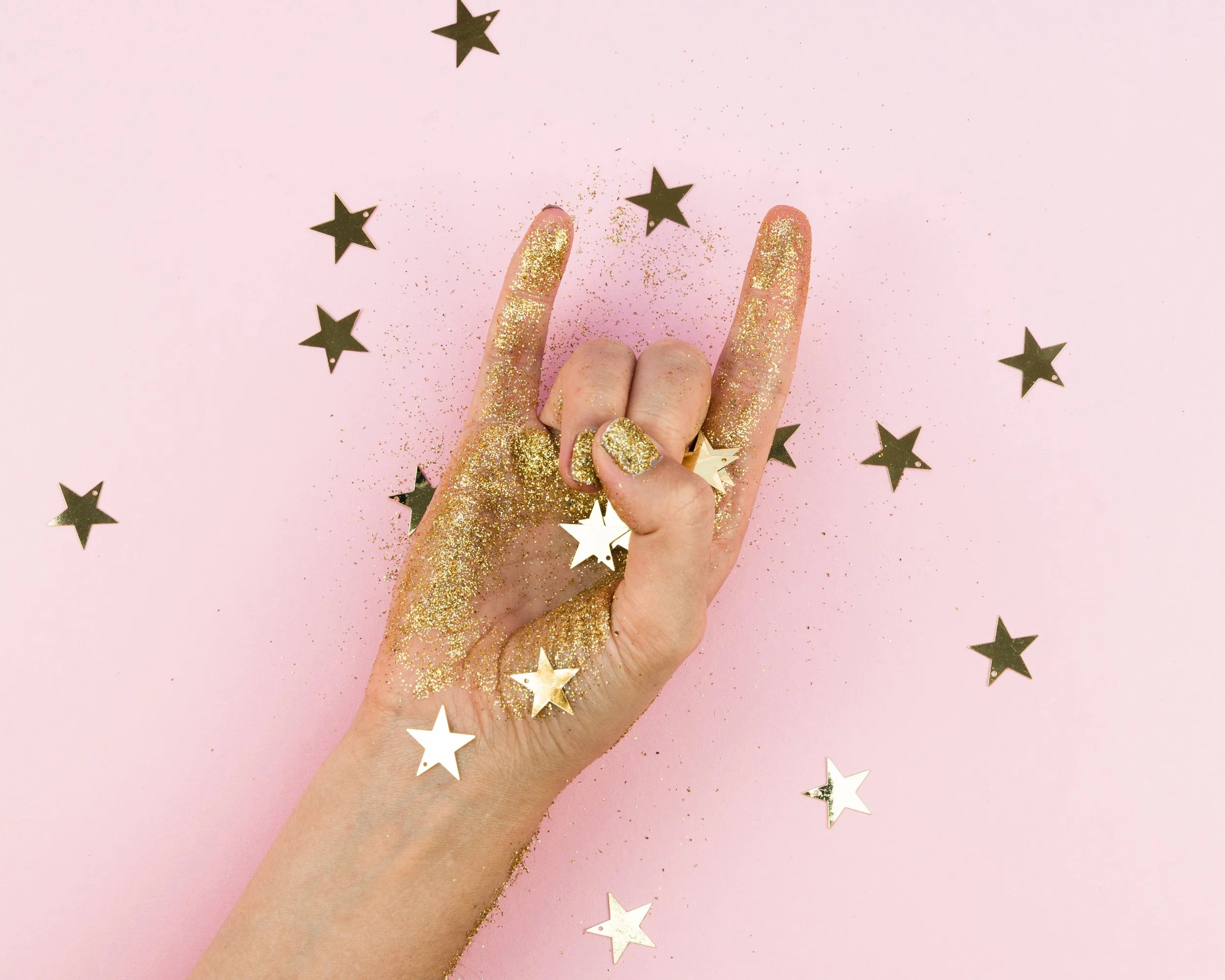 A hand with gold glitter on fingers and thumb painted gold, holding a few gold star-shaped confetti pieces, against a pink background with scattered black and white star-shaped confetti.