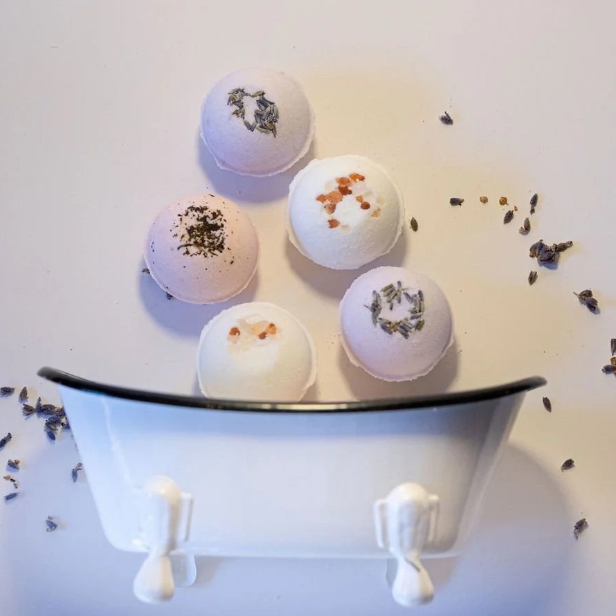 Five bath bombs with different colors and dried herbs on top, placed inside a white container with a black rim.