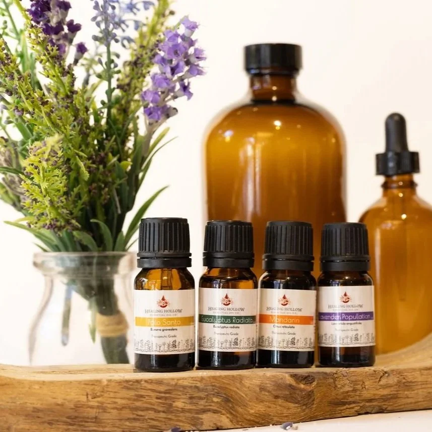 A display of essential oils in small amber glass bottles, with a larger amber bottle and a dropper bottle behind them, alongside a vase of lavender and greenery on a wooden surface.
