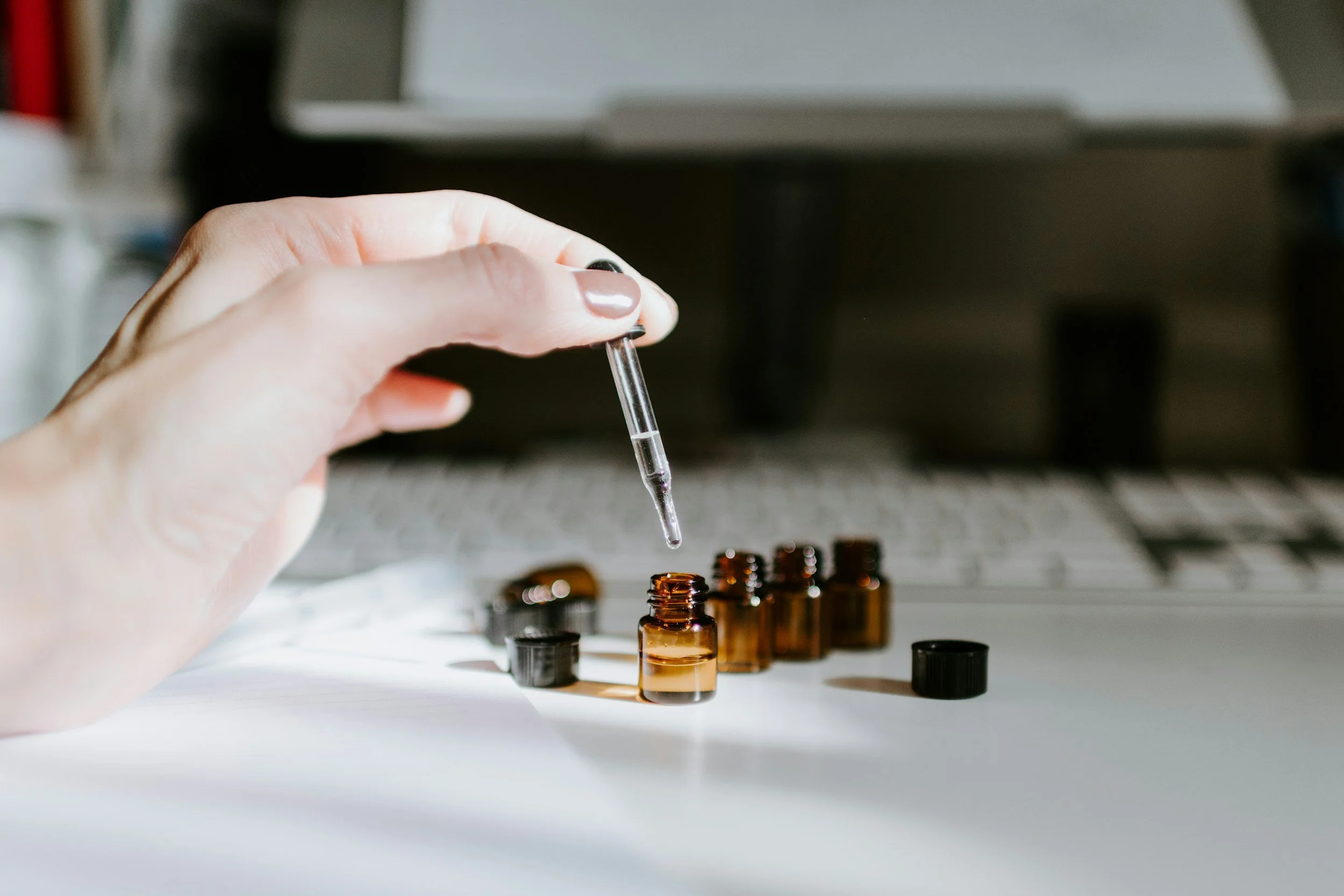 A person’s hand with painted nails holding a glass dropper, transferring liquid into small amber glass bottles on a white surface with a computer keyboard and monitor in the background.