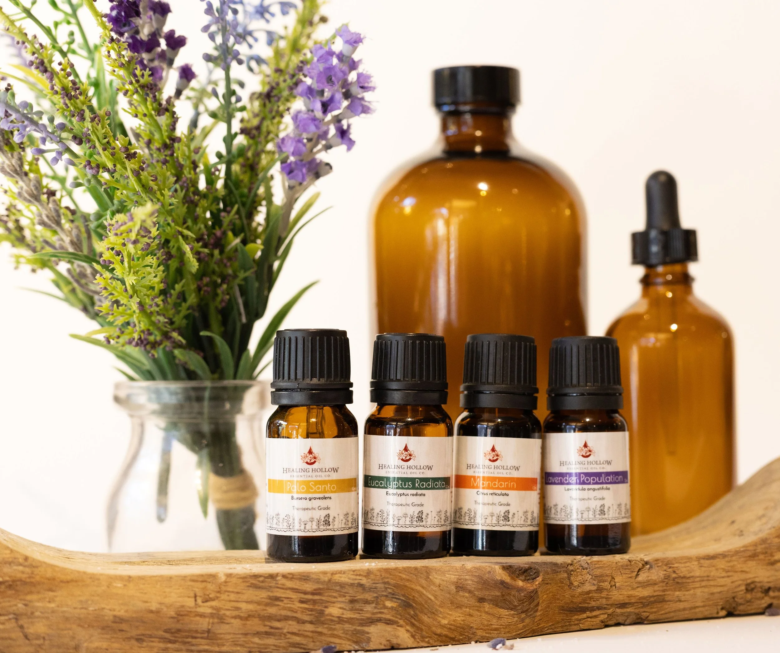 A display of four small Amber bottles labeled with essential oils, a large amber bottle, a smaller amber dropper bottle, and a vase of purple and green leafy flowers on a wooden surface.