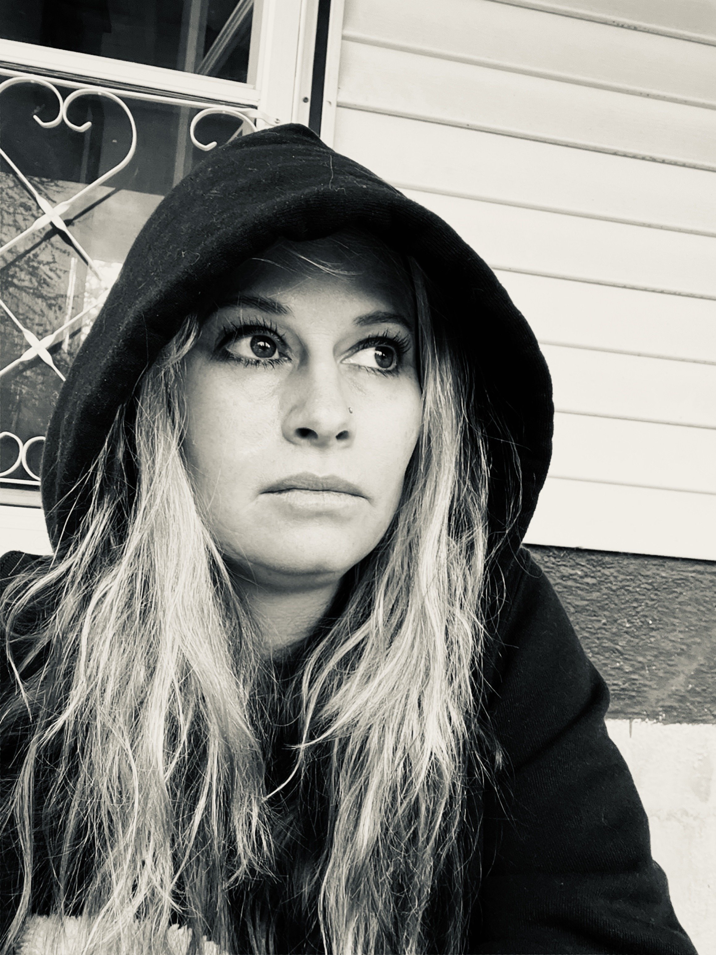 A woman with long blonde hair wearing a black hoodie, looking off to the side in front of a house with horizontal siding and a window with decorative metalwork.