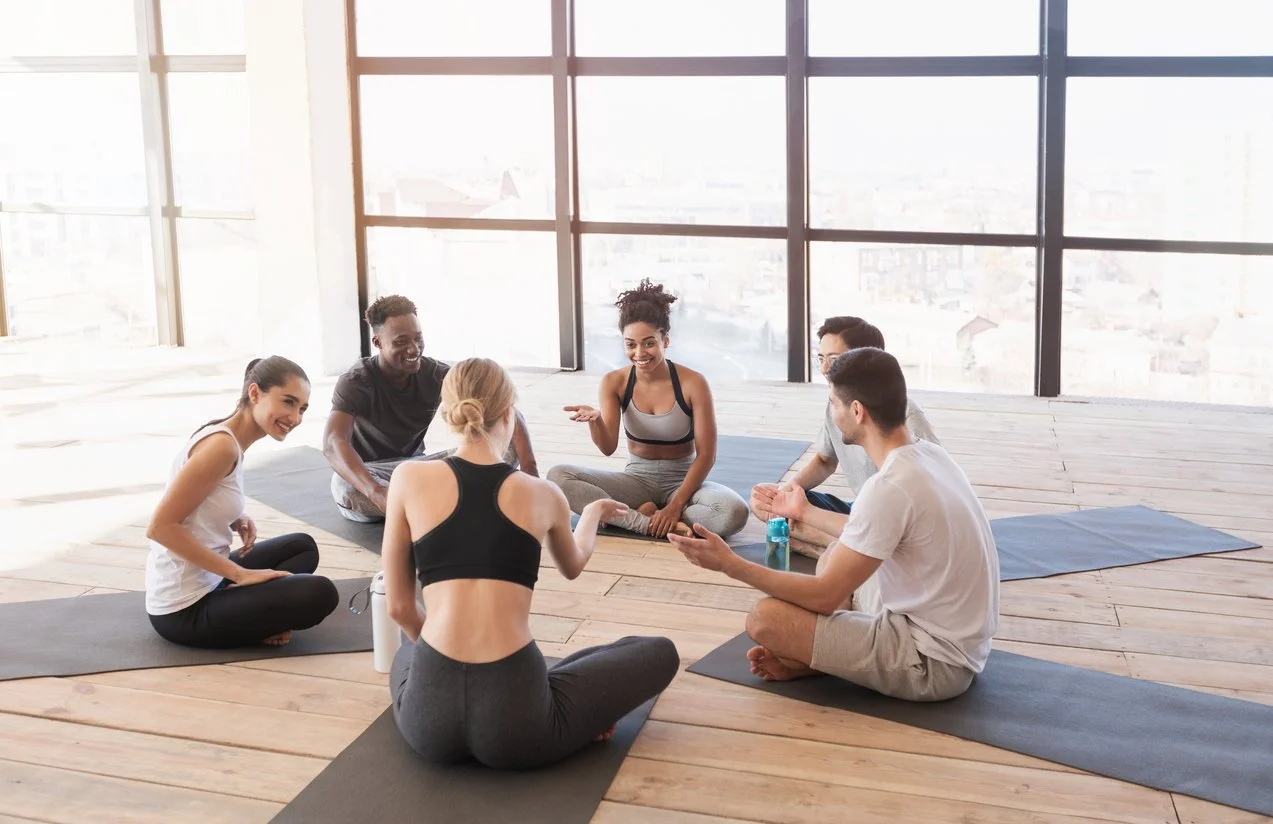 Group of six people sitting on yoga mats in a circle, engaging in a conversation in a bright room with large windows.