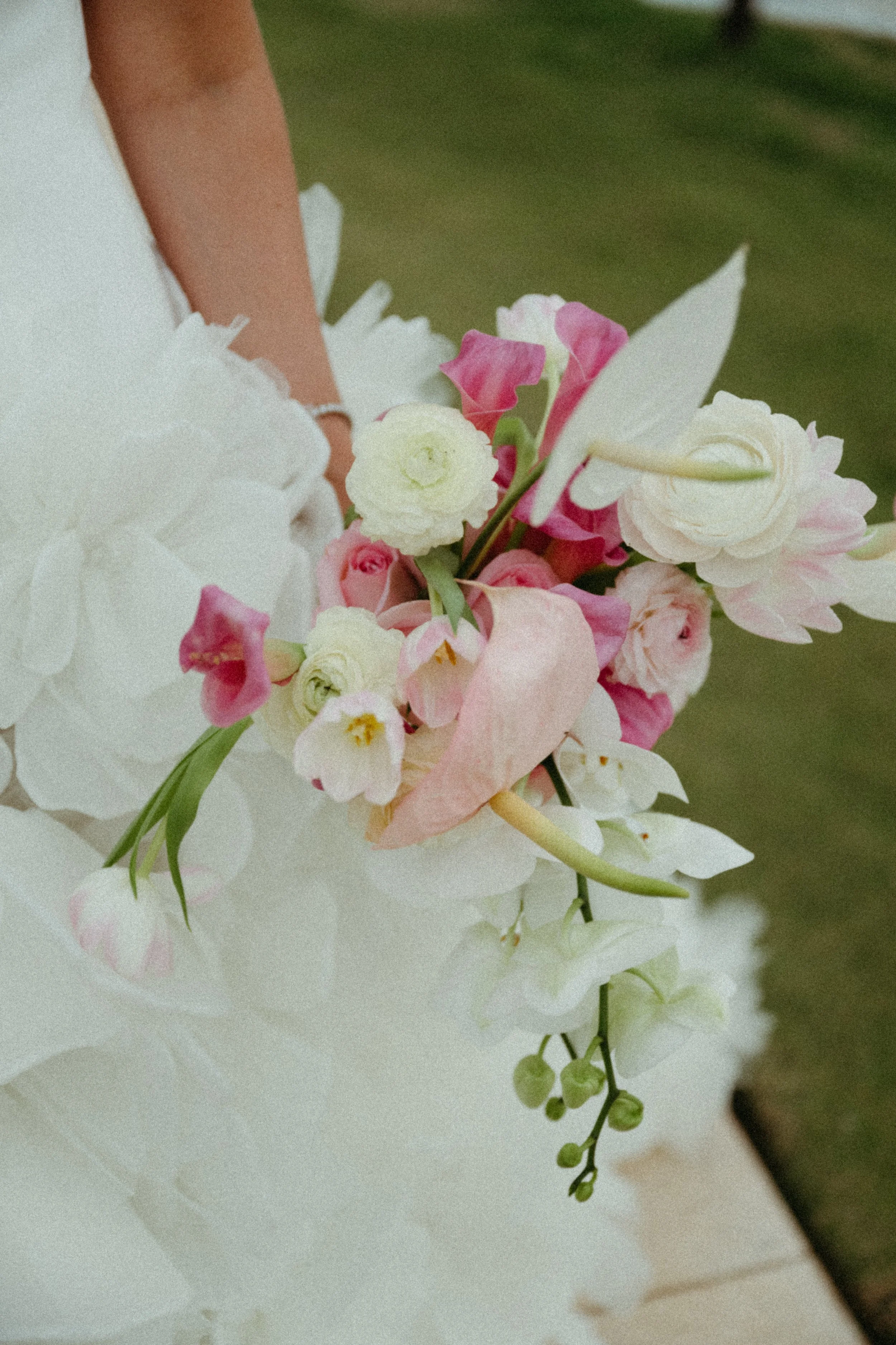 Close-up of a bride holding a pastel-colored flower bouquet with pink and white flowers, and a white dress, outdoors.