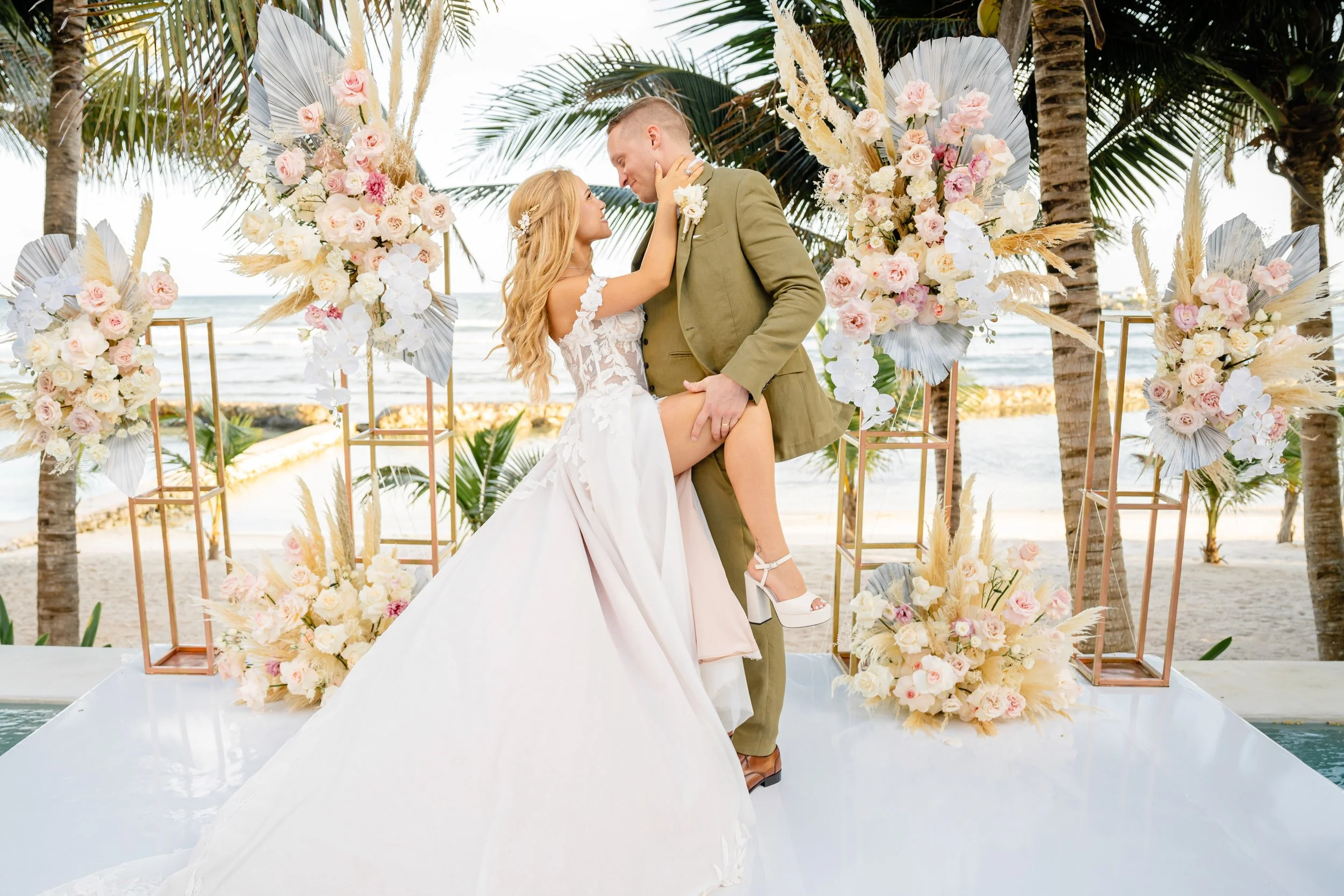 A bride and groom in wedding attire on a beach, embracing and smiling. The scene features floral arrangements with pink and white flowers and greenery, palm trees, and ocean in the background.