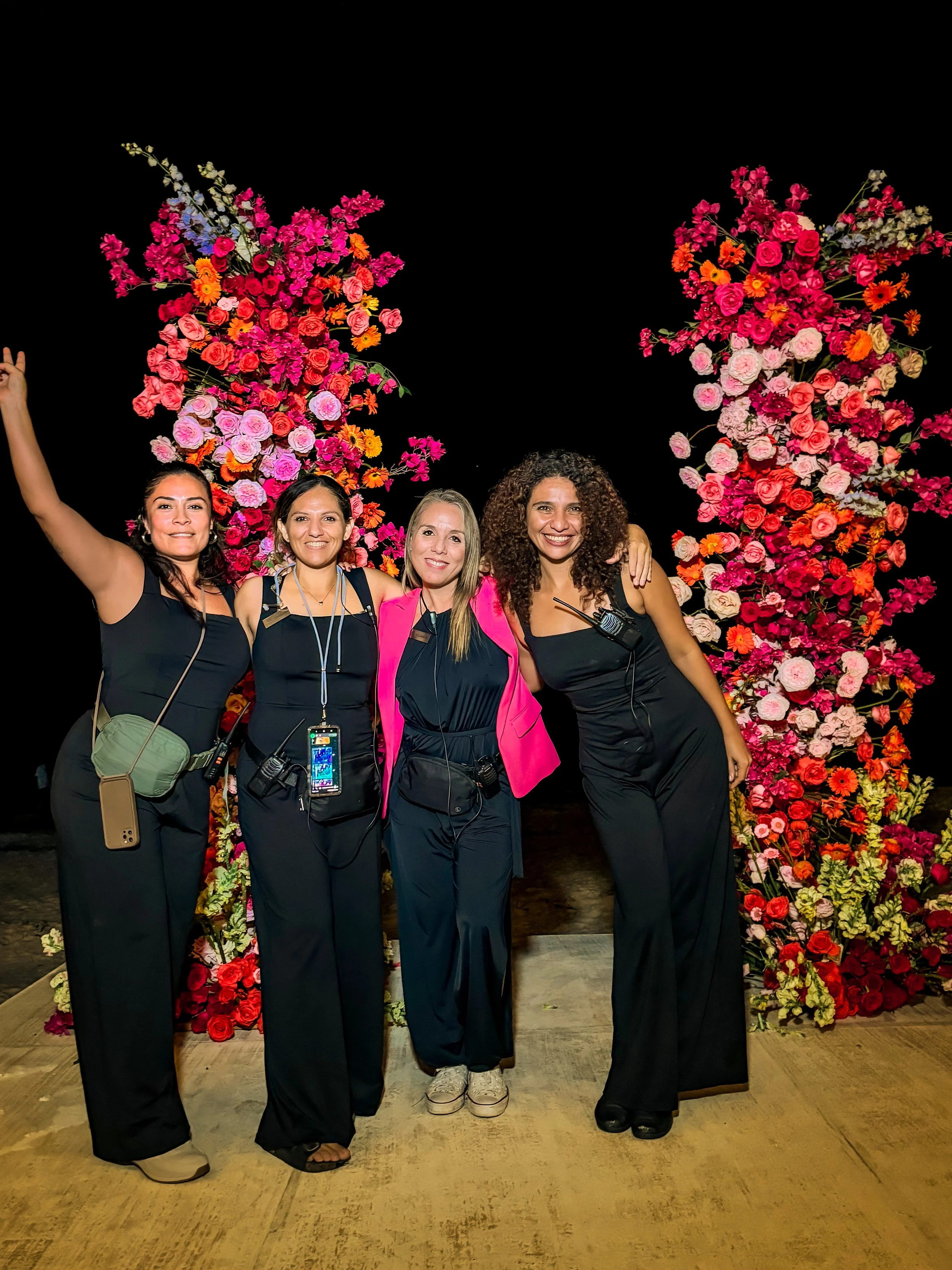Four women standing in front of vibrant flower arrangements at a night event, smiling and posing for the photo.