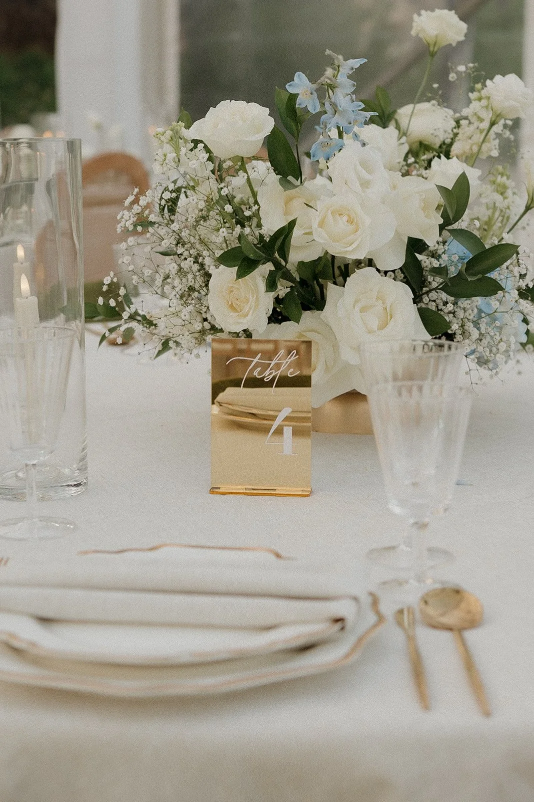 Elegant table centerpiece with white roses, blue flowers, greenery and baby's breath; table number card reading 'Table 4'; set glasses, white plates, gold utensils, and candles in glass holders on a white tablecloth.