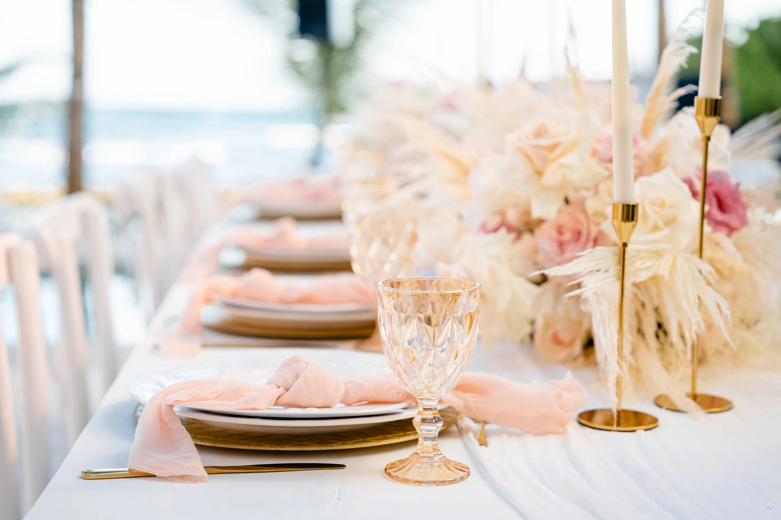 Elegant wedding table setting with gold-rimmed plates, pink napkins, crystal glasses, and a floral centerpiece with soft pink and white flowers, gold candlesticks, and candles.