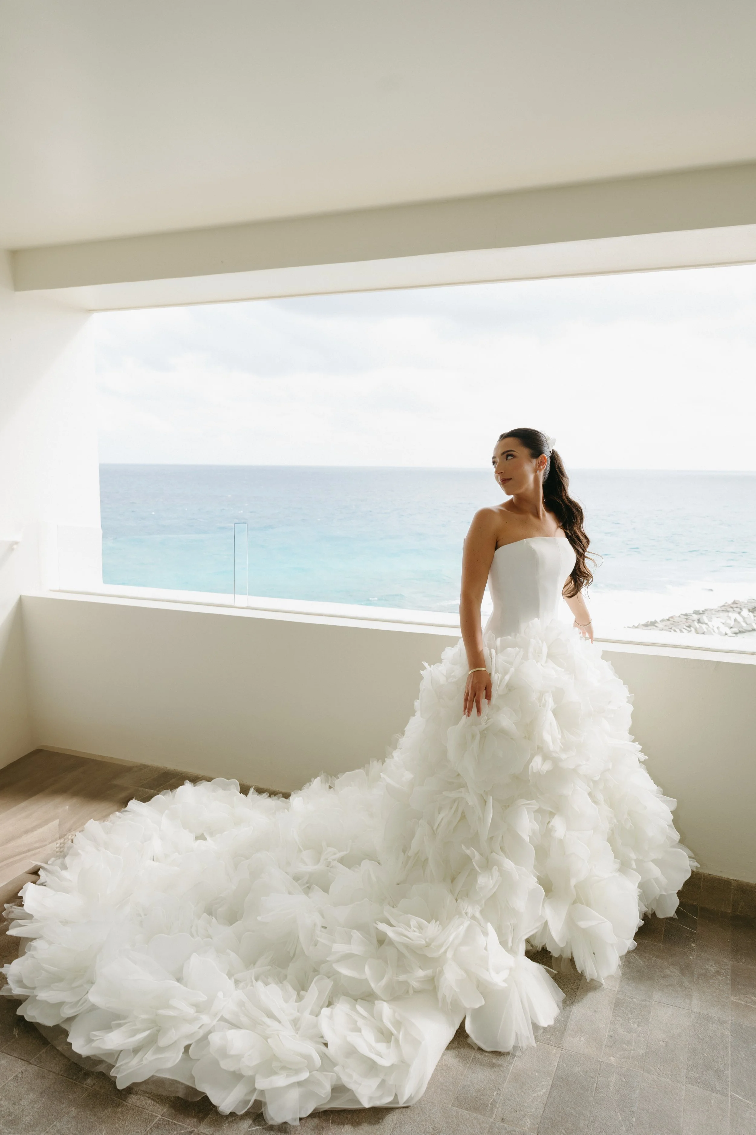 A bride in a strapless white wedding gown with a voluminous, ruffled train standing inside near a large window overlooking the ocean.