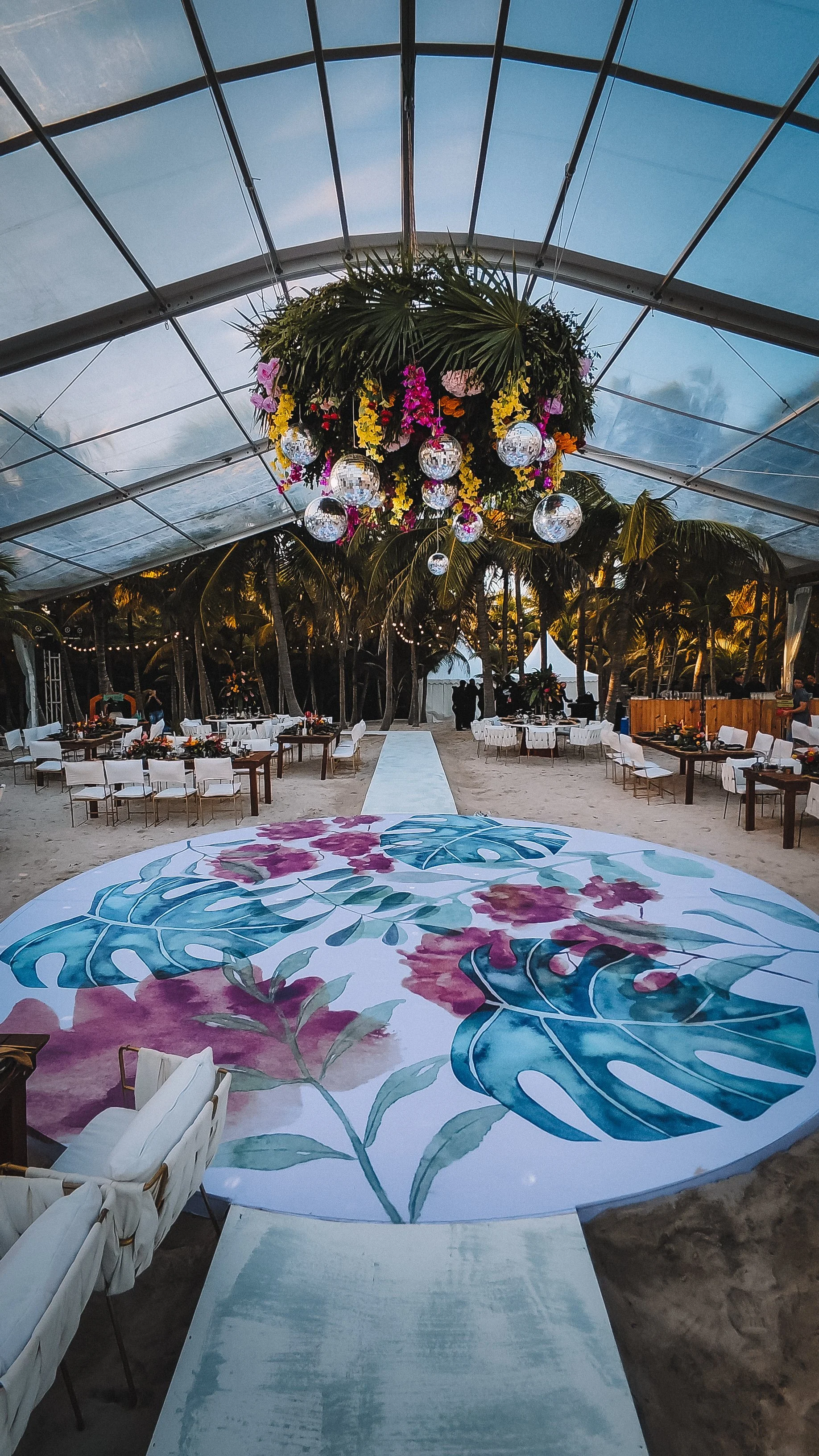 Wedding or event setup on a beach with a floral and disco ball chandelier, decorated tables, and a painted floral walkway leading to the background with palm trees and a tent.