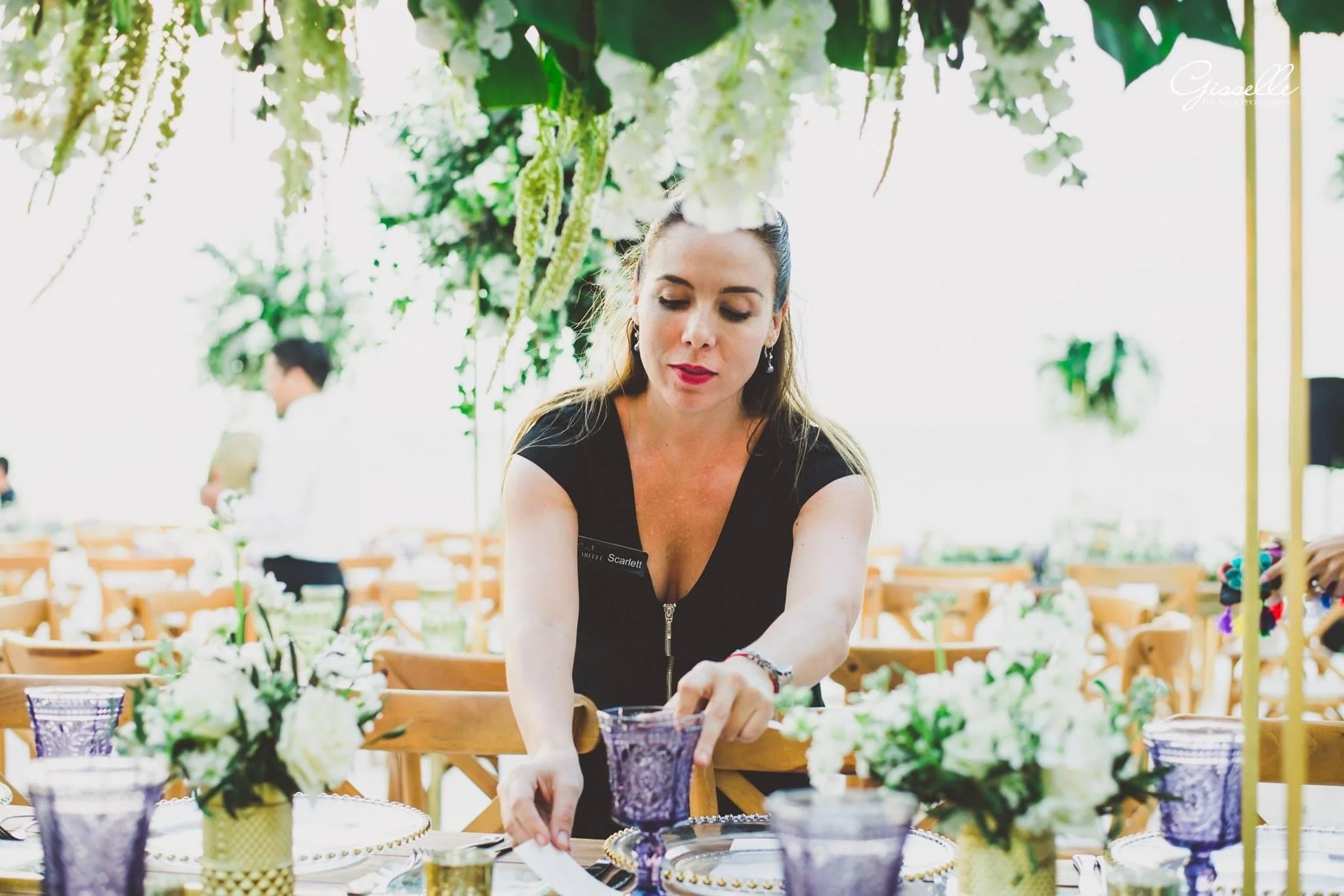 A woman preparing a table setting at a decorated event with floral arrangements and purple glassware, with greenery overhead and background figures.