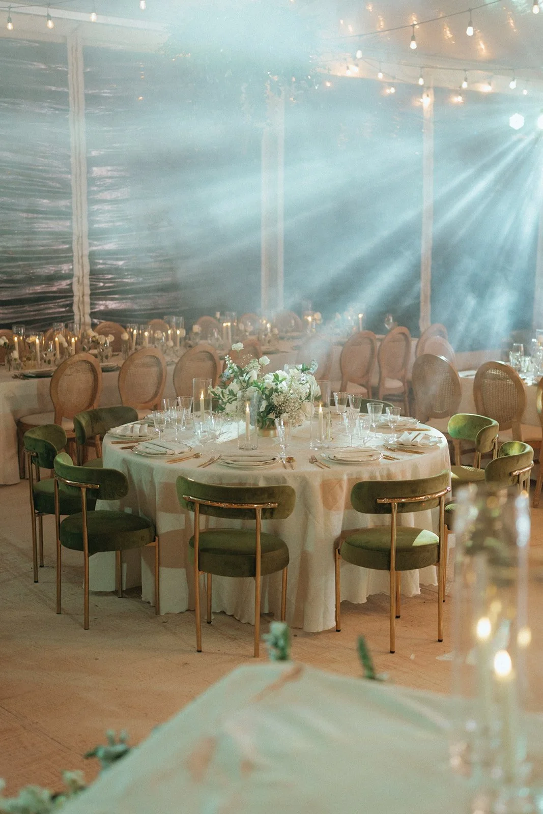 Elegant event space with round table set for a formal dinner, decorated with a floral centerpiece, candles, and tableware, with string lights and chairs arranged around the tables under a tent.