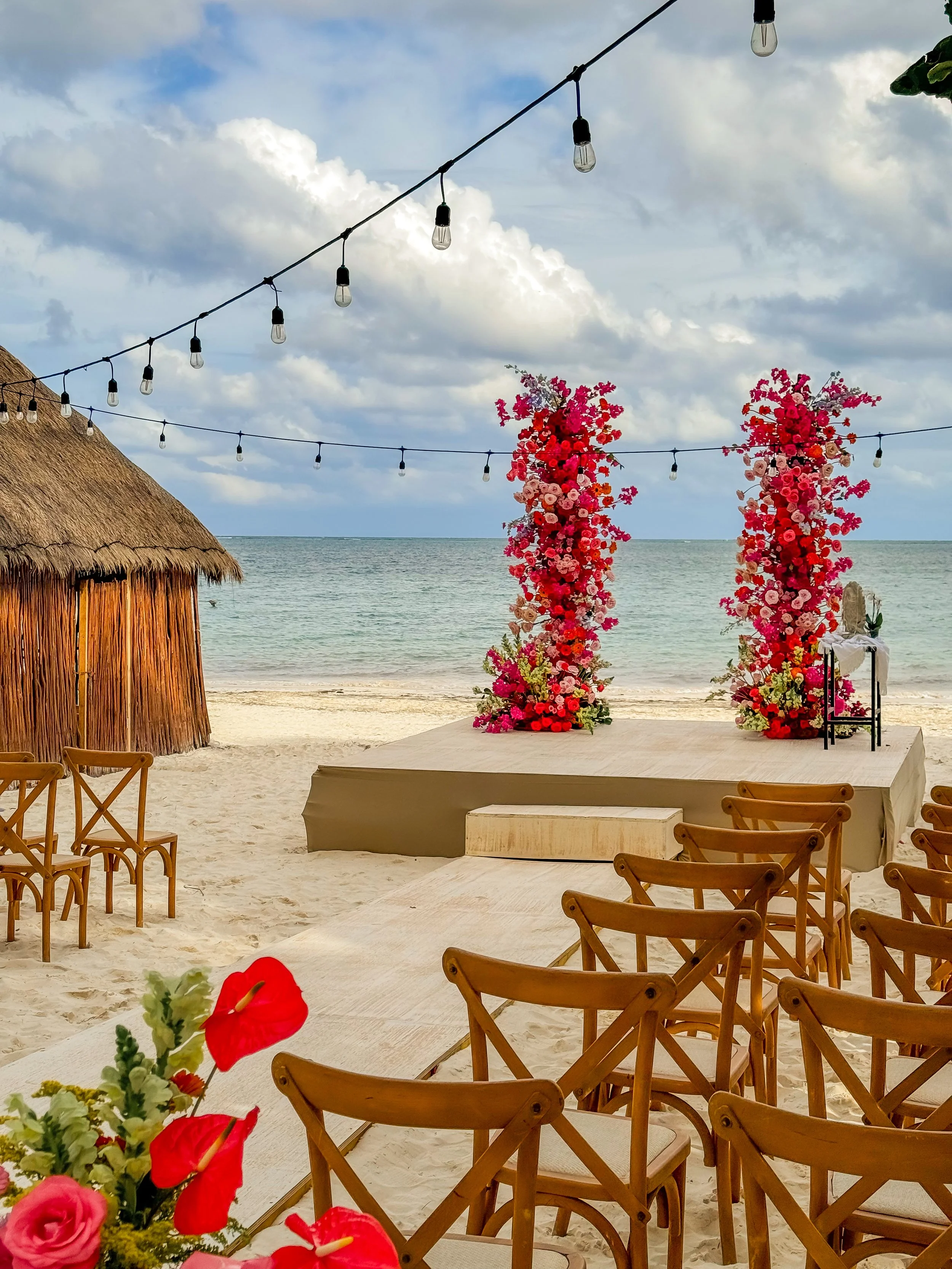 Beach setup for a wedding with floral arch, chairs, and a table, under string lights, by the sea with a thatched hut nearby.