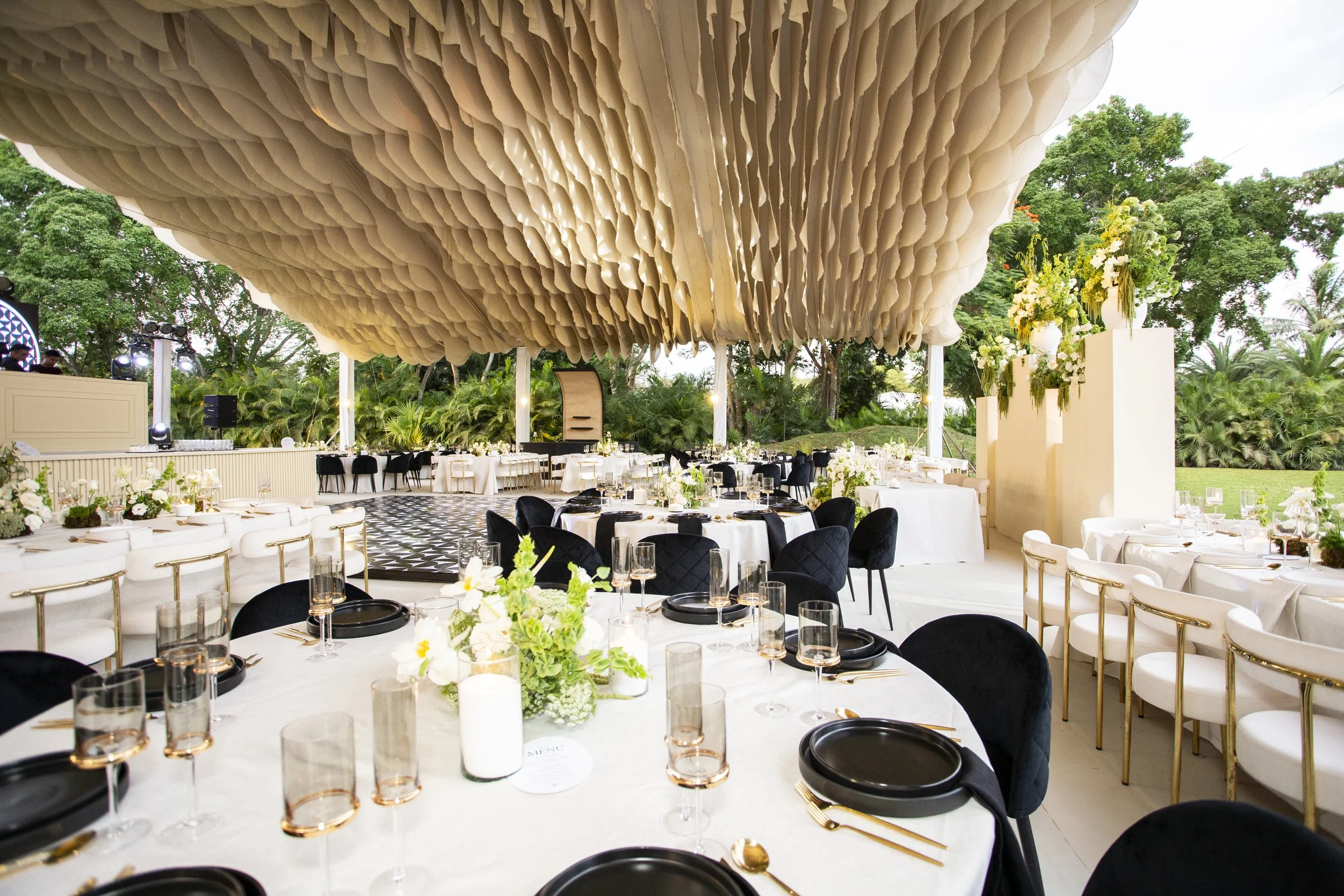 Elegant outdoor event at a Yucatan Hacienda  with round tables set with black and gold tableware, white floral centerpieces, and black and white chairs beneath a large beige fabric ceiling installation, surrounded by greenery and trees.