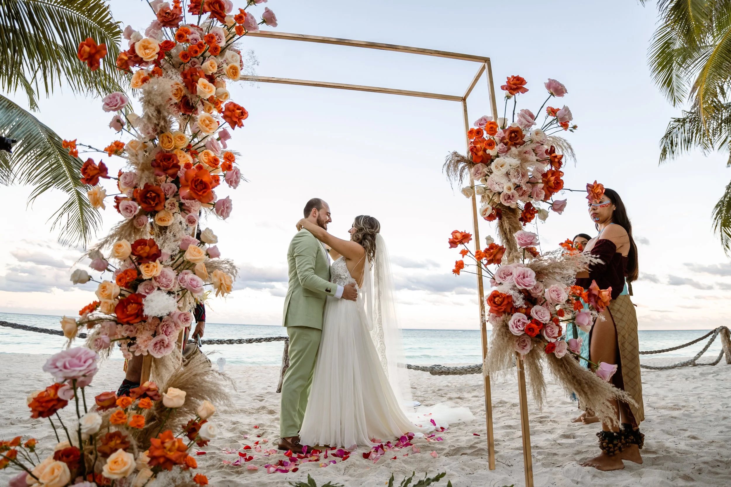 Beach wedding ceremony with a couple exchanging vows under a floral arch, surrounded by palm trees, on the sand by the ocean at sunset.